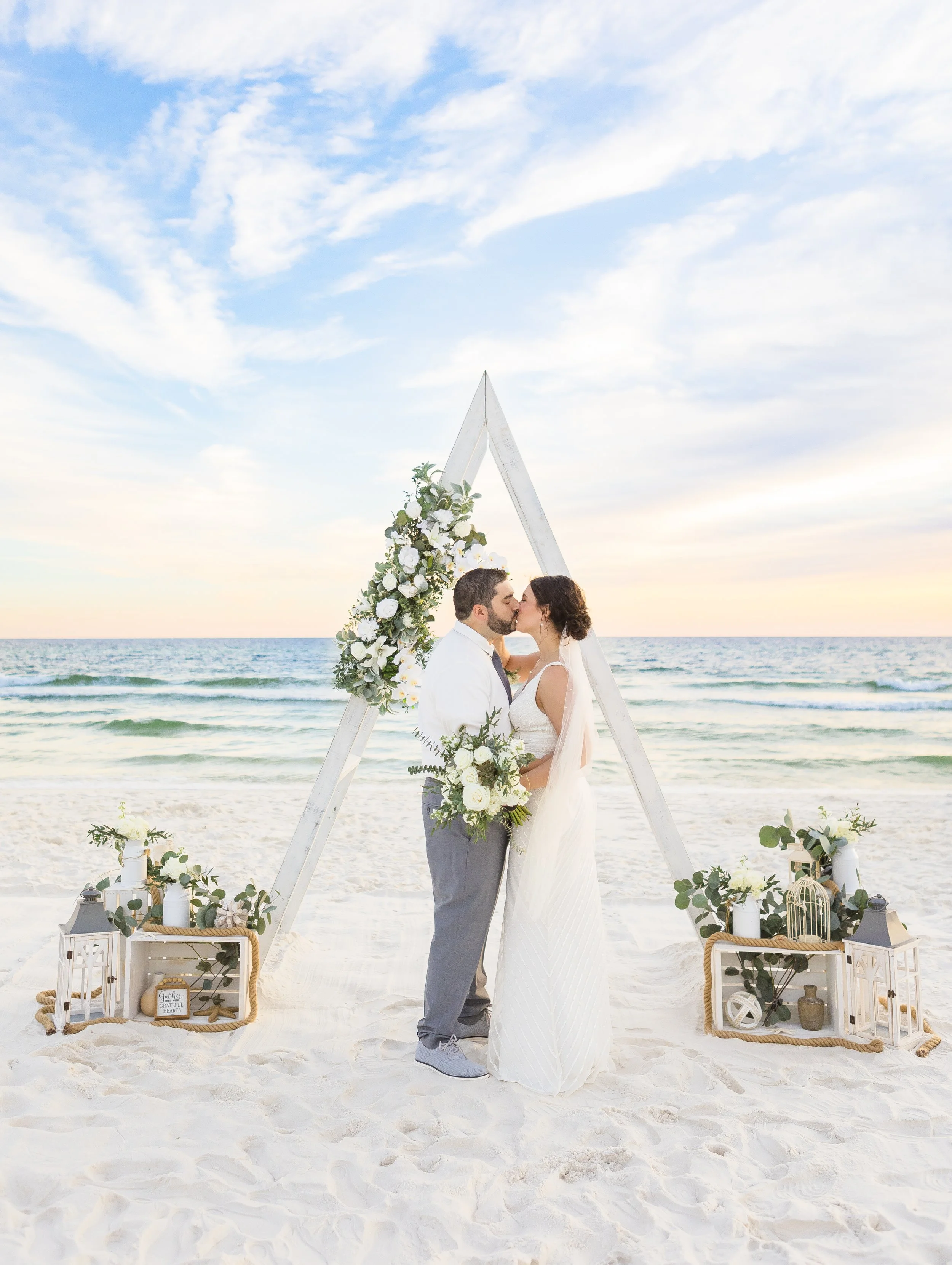 A couple in wedding attire sharing a kiss on a beach during their wedding ceremony, with an arch decorated with flowers and greenery, and decorative lanterns and signs on the sand.