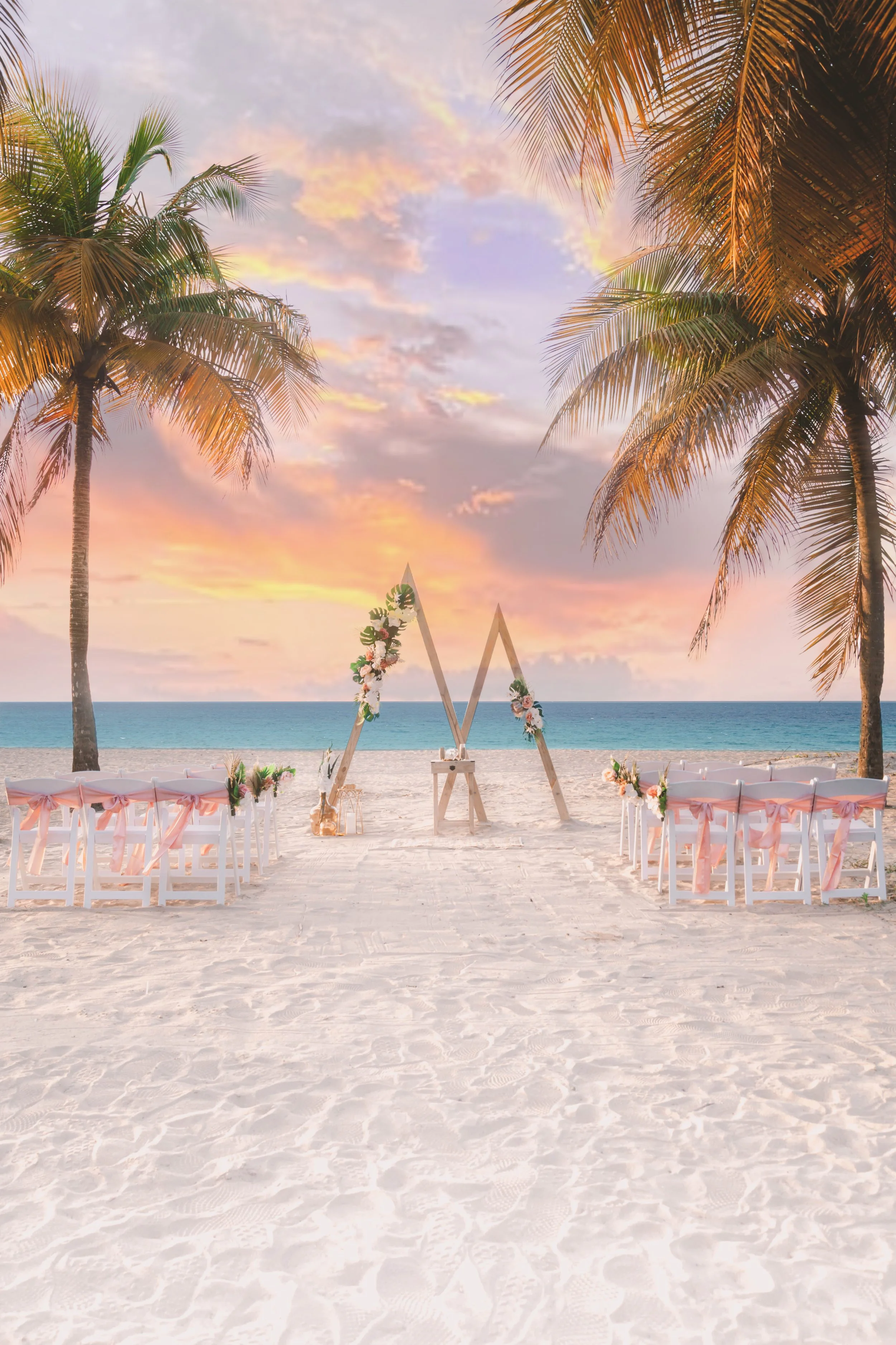 Beachside wedding setup with chairs decorated with pink ribbons, an arch with flowers, two tall palm trees, and a sunset sky.