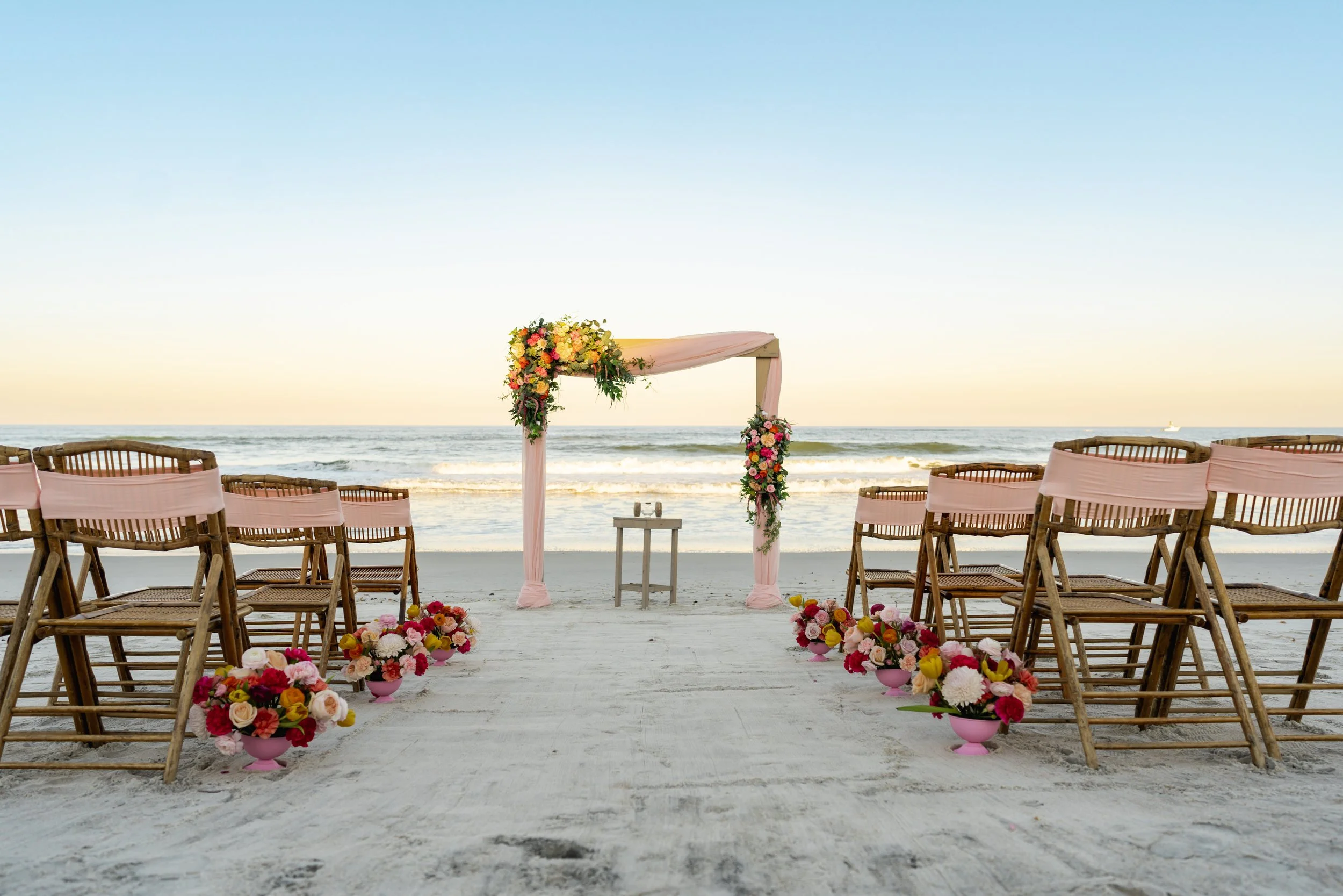 Beach wedding ceremony setup with pink floral decorations and wooden chairs facing the ocean at sunset.