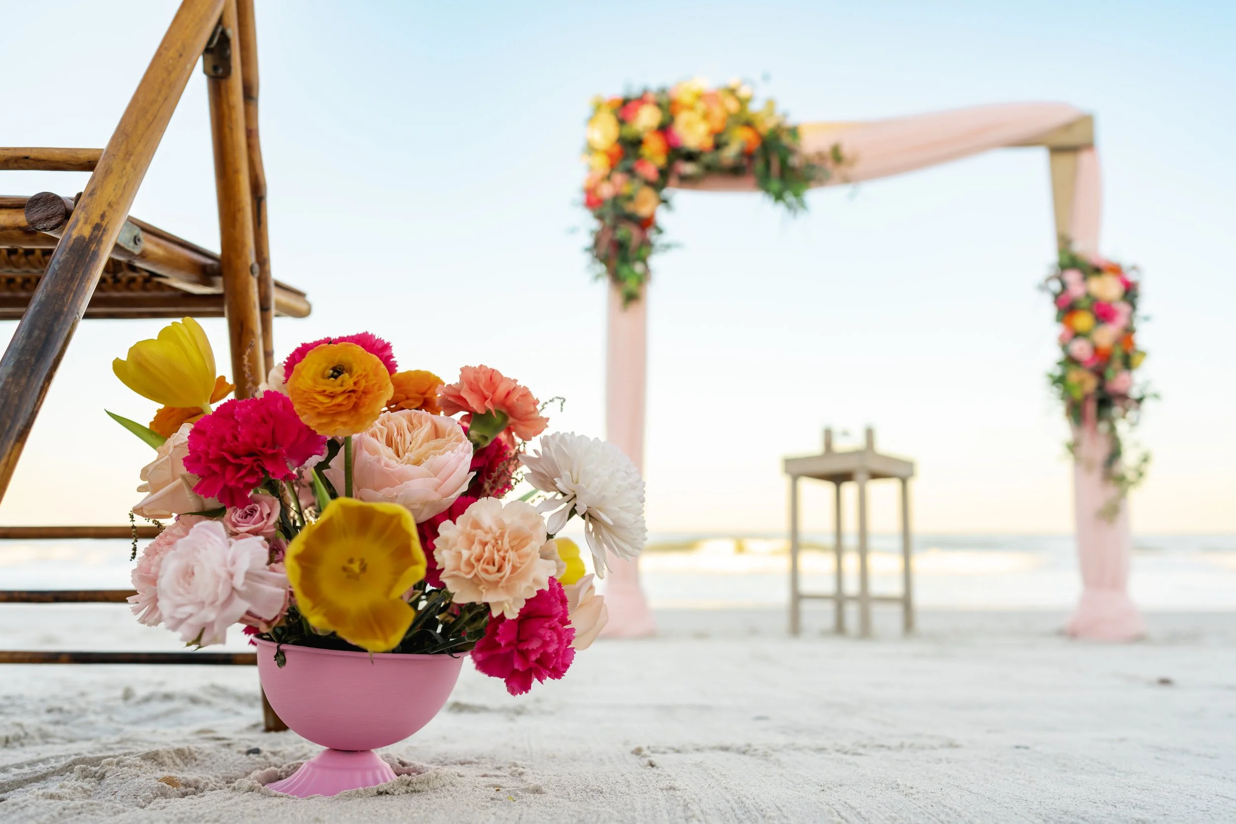 Beach wedding setup with a pink flower arrangement in the foreground, an arch decorated with flowers, and a small table against the ocean backdrop.