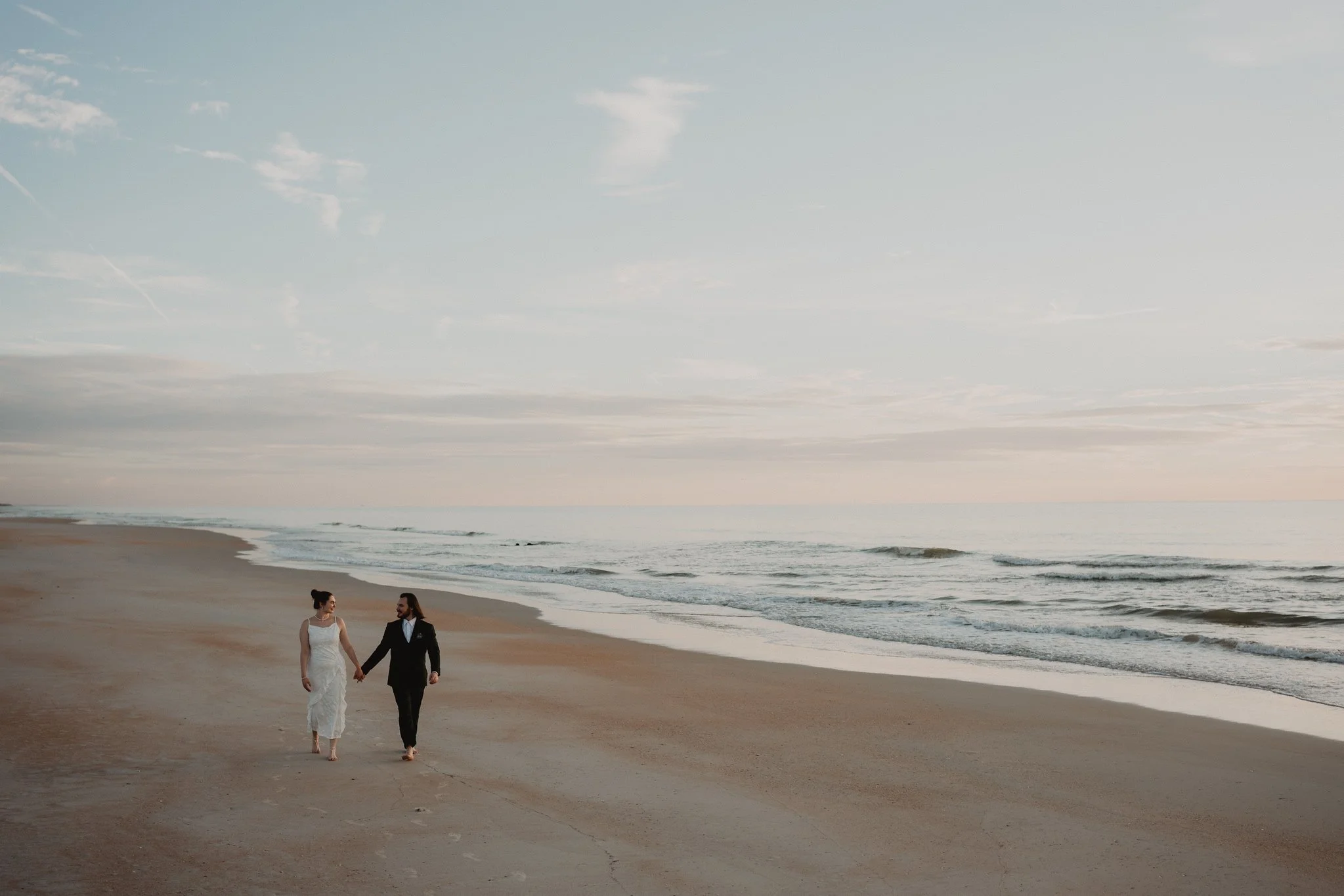 A couple in wedding attire holding hands and walking along a sandy beach with the ocean and a cloudy sky in the background.