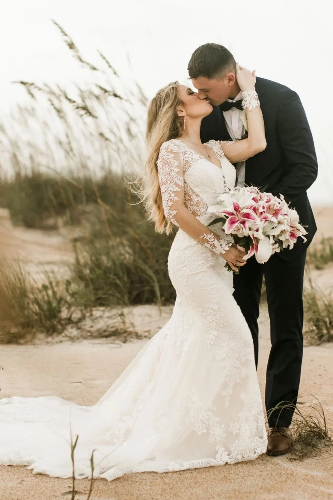 A bride and groom kissing on a sandy beach. The bride is wearing a long white lace wedding dress and holding a bouquet of pink and white lilies. The groom is dressed in a black tuxedo. There are tall grasses in the background.