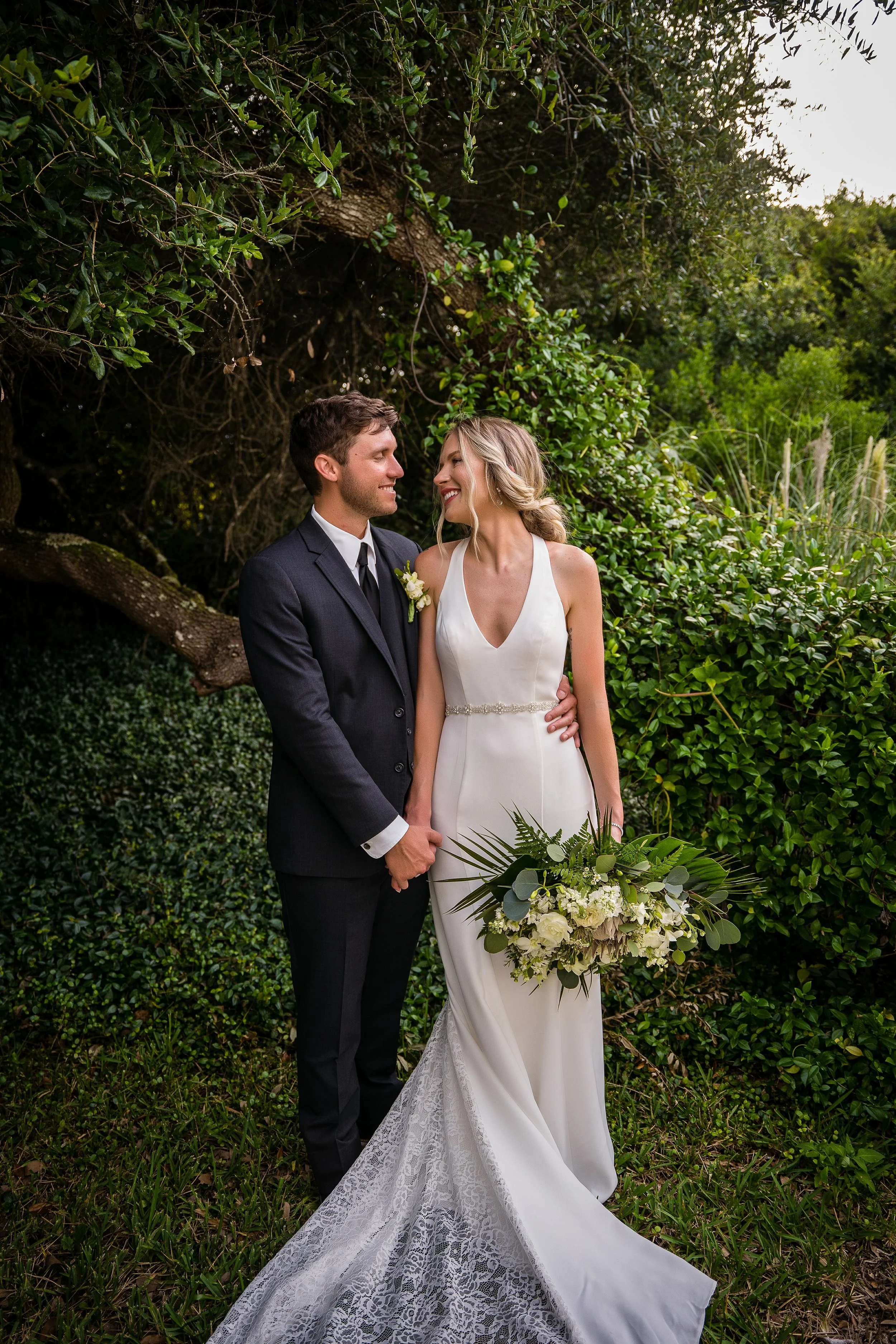 A newlywed couple standing outdoors, holding hands, surrounded by lush green foliage. The bride wears a white satin wedding gown with lace train and holds a bouquet of white flowers and green leaves. The groom wears a dark suit with a boutonniere. Th