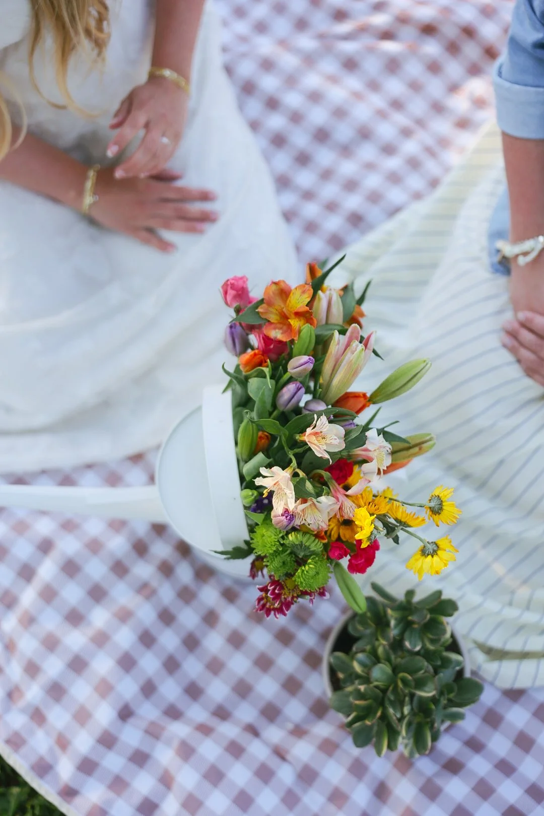 two women sitting around a watering can with flowers in it next to a succulent plant