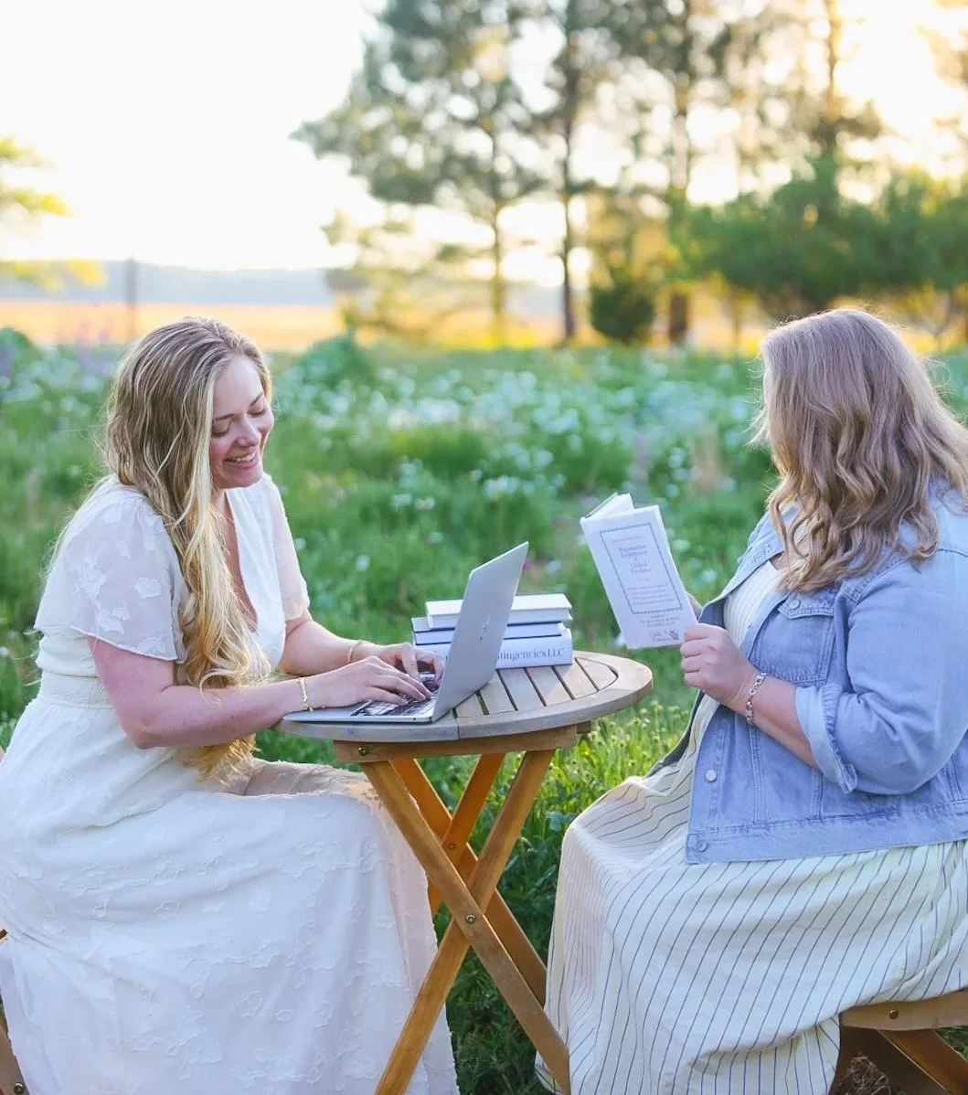 Two women sitting at a small wooden table outdoors on a grassy field, working on laptops and reading books, surrounded by trees and greenery during sunset.