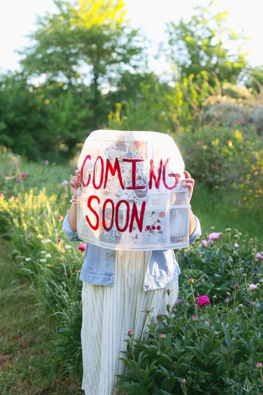 A person holding a sign that says 'COMING SOON...' written in red letters covering their face. The person is standing outdoors in a garden with green bushes and pink flowers, with trees and sunlight in the background.