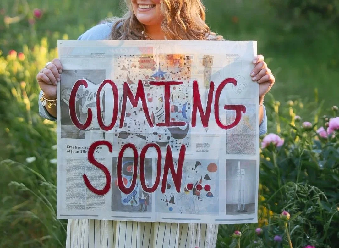 A woman holding a sign that says 'COMING SOON' written in red marker, standing outdoors in a garden with pink flowers.