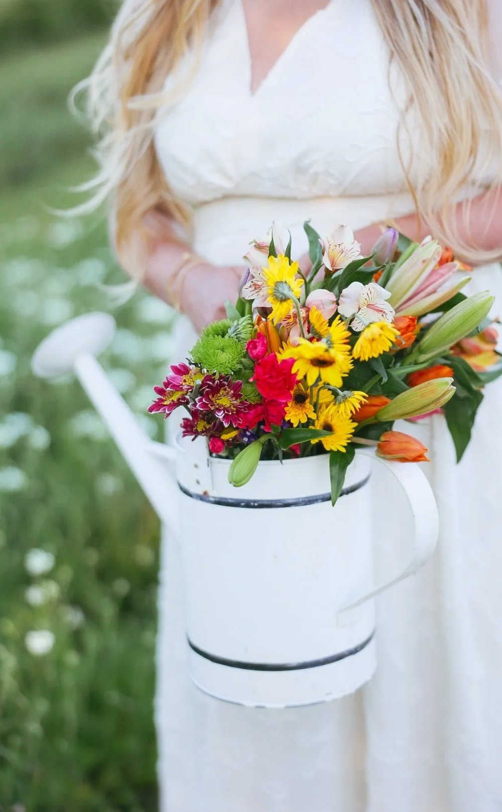 A woman in a white dress holding a white watering can filled with colorful flowers, standing outdoors in a grassy area.