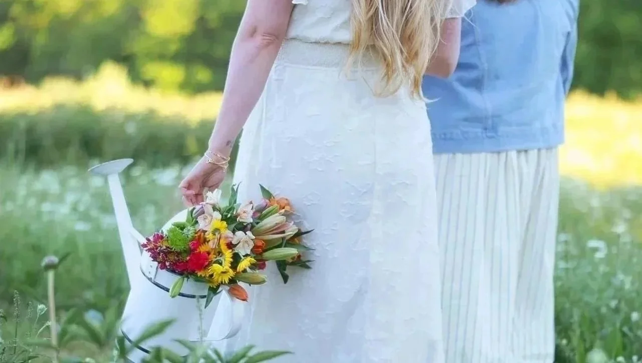A woman holding a white watering can filled with colorful flowers, standing outdoors near a pond with green trees in the background.
