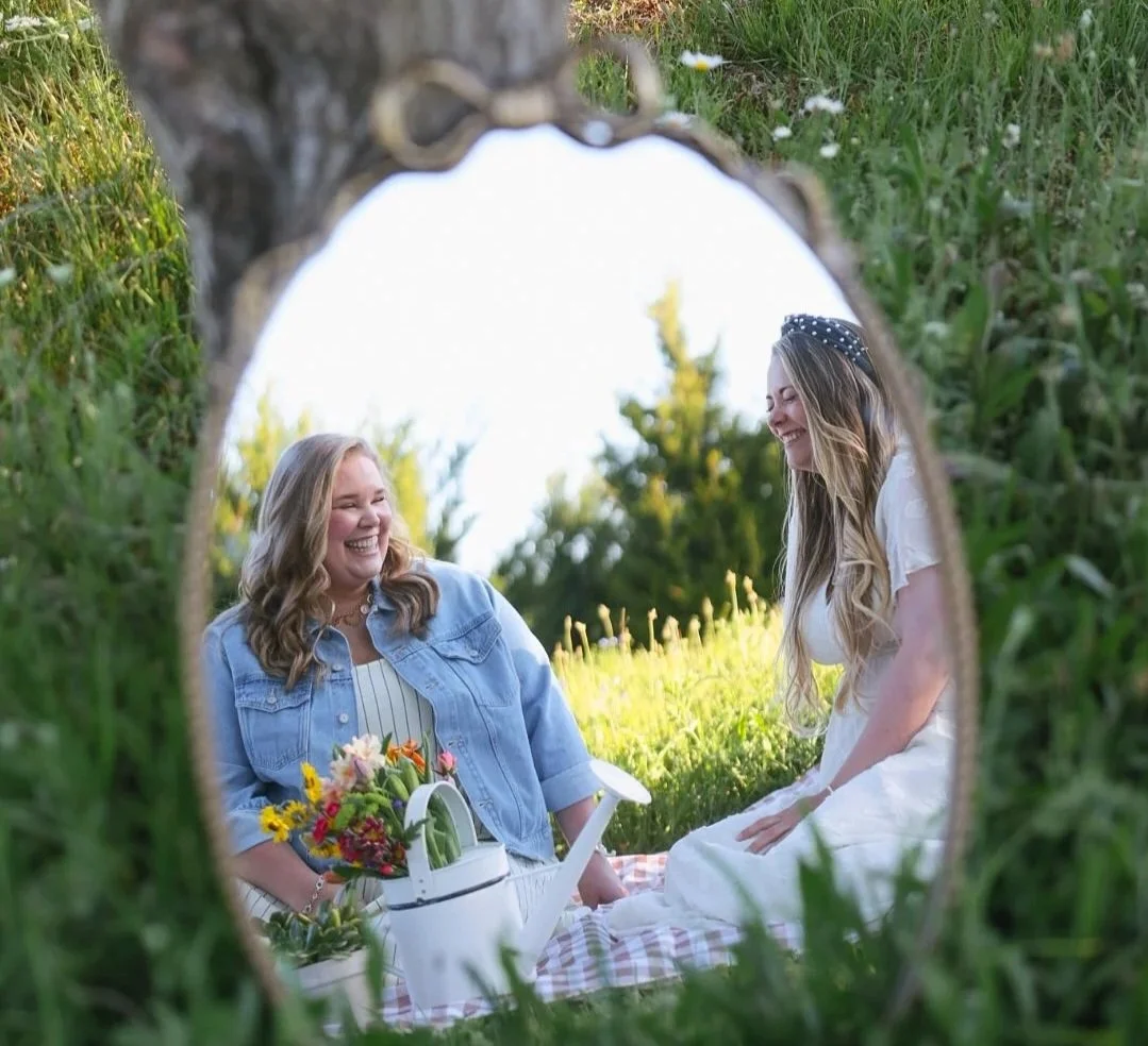 Two women having a picnic outdoors, viewed through a tree hollow. One woman wears a denim jacket and the other wears a white dress and a headband. They are smiling and laughing, with flowers and a watering can on a blanket.