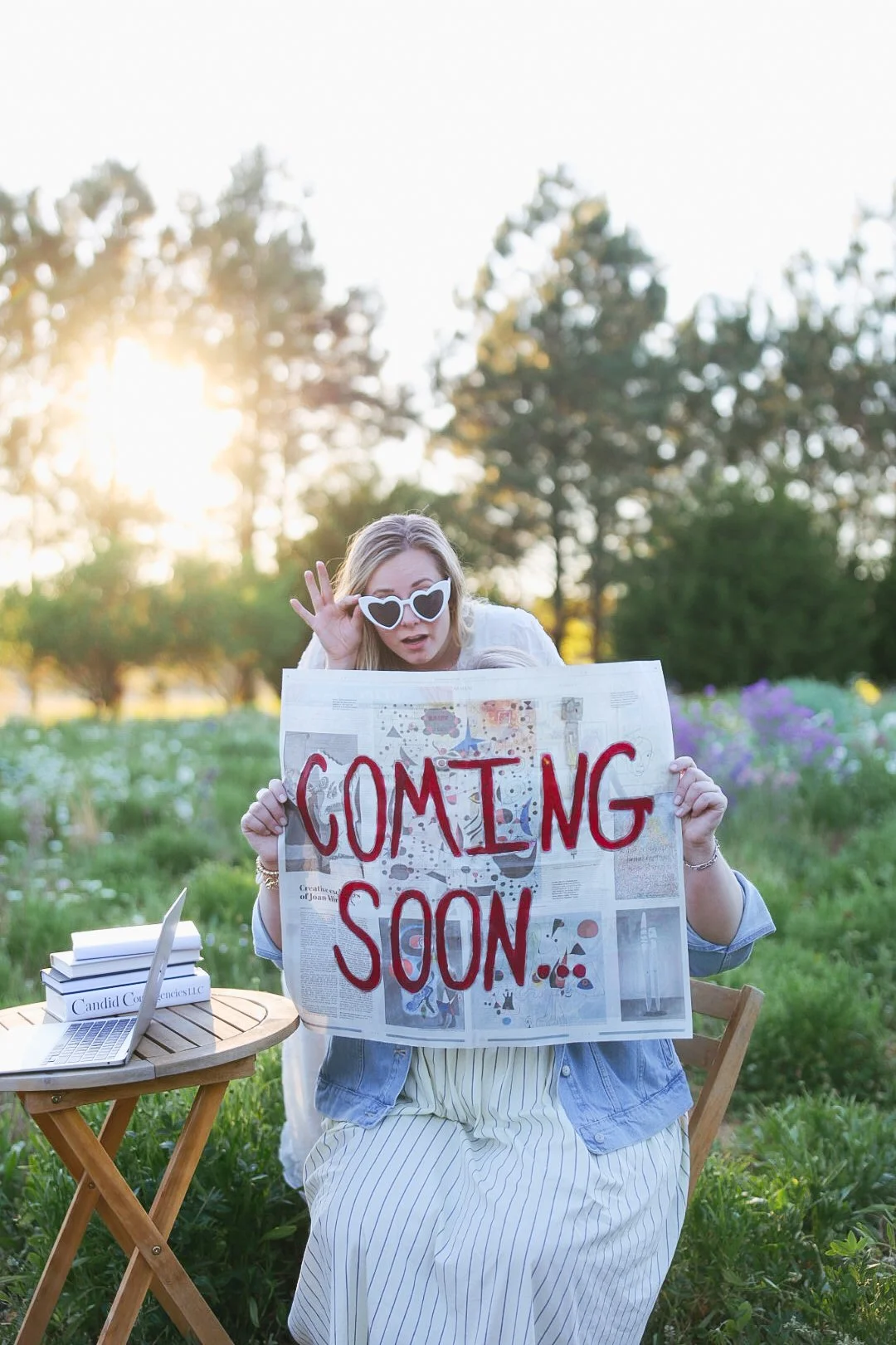 Woman with blonde hair wearing sunglasses and a denim jacket, holding a sign that says 'Coming Soon' in red letters, sitting outdoors in a park with trees and sunlight in the background.