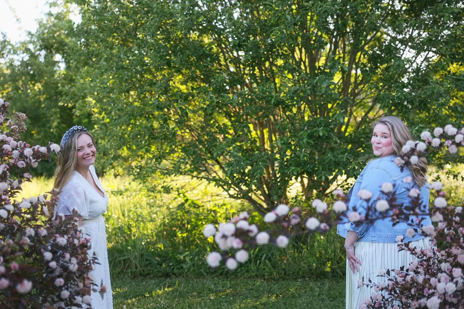 Two women standing outdoors among flowering bushes and green trees, smiling and looking at the camera during late afternoon.