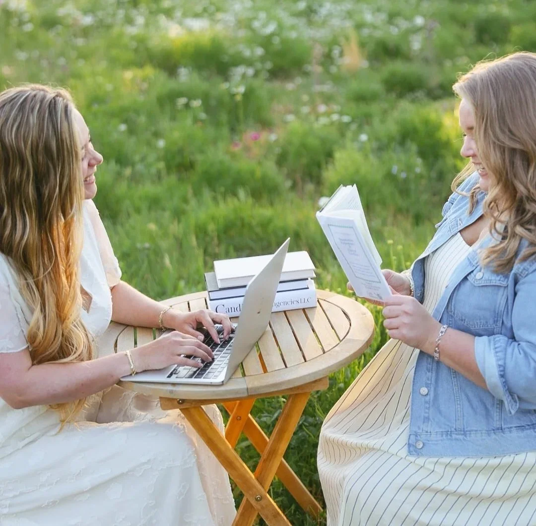 Two women smiling while sitting at a small table outside using a laptop and reading a book.