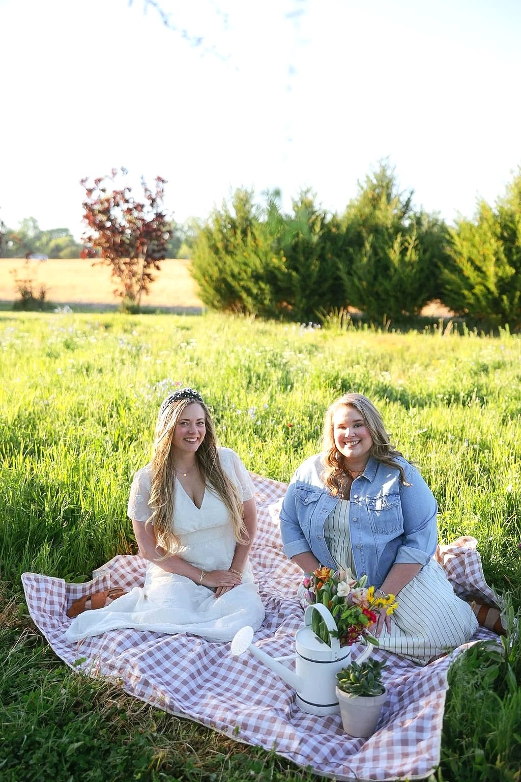 Two women enjoying a picnic on a checkered blanket in a grassy field with trees in the background, bright sunlight, and a clear sky.