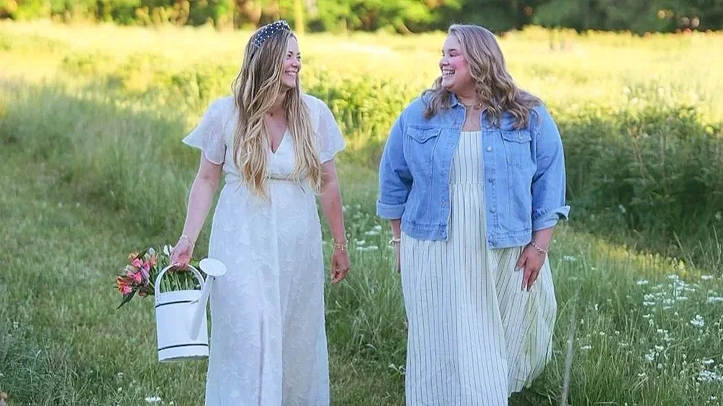 Two women laughing while while walking through a field and holding a watering can with flowers in it.