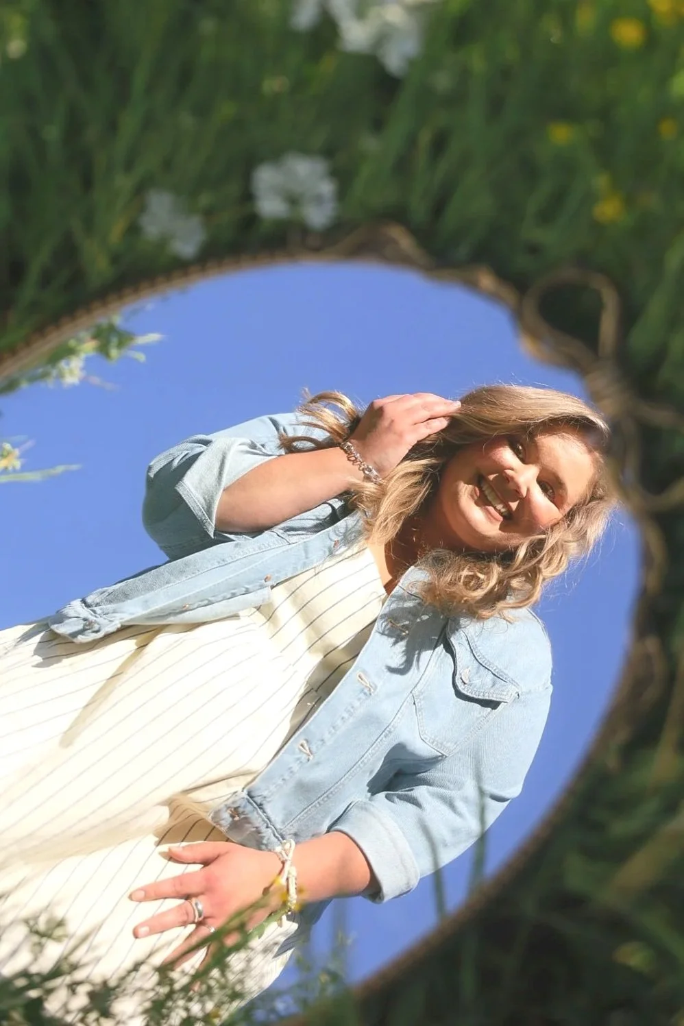 A woman with blonde curly hair smiling, standing outdoors, viewed from below through a circular frame of tree branches and leaves, with a clear blue sky in the background.