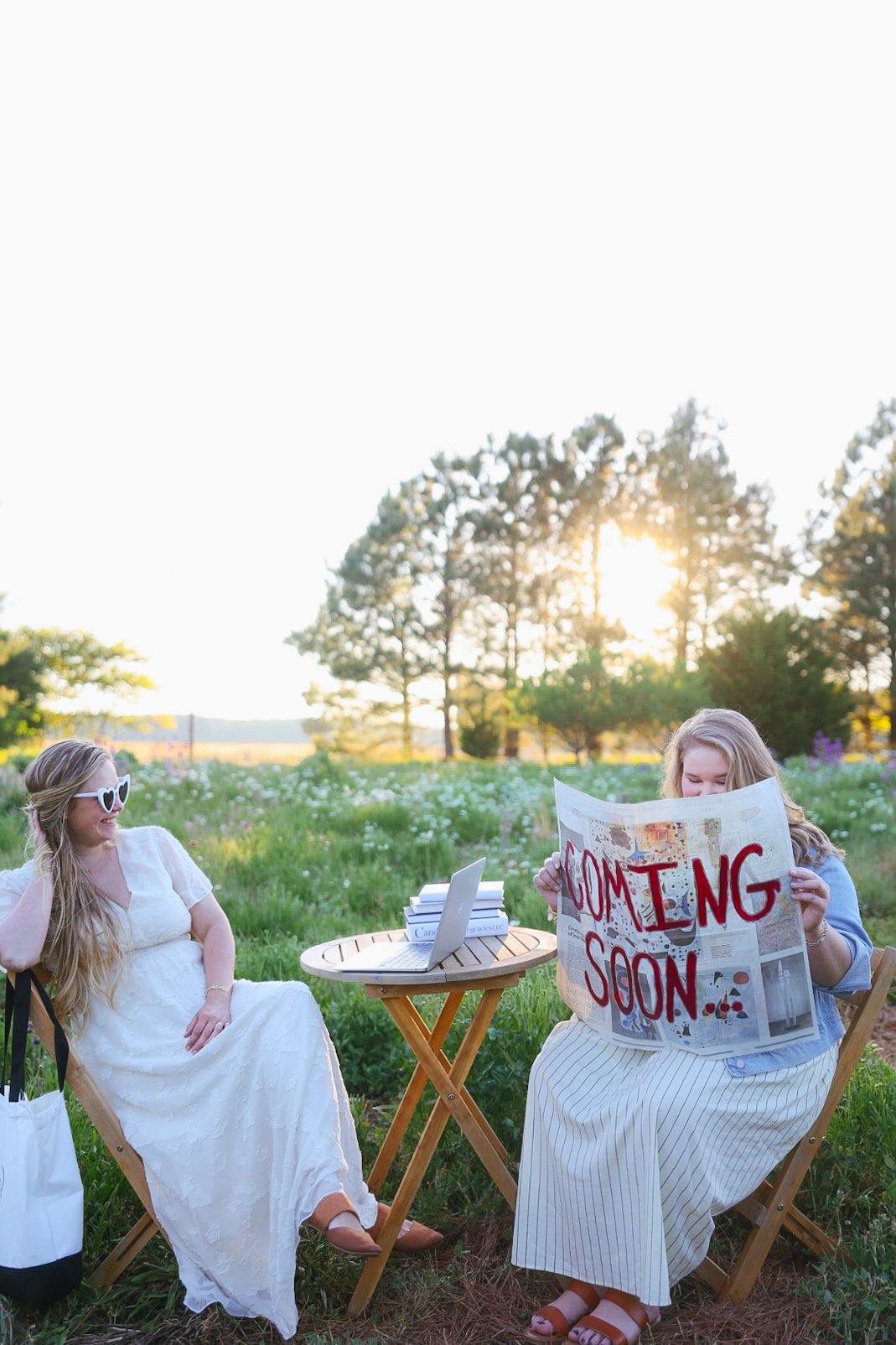 Two women sitting outdoors at a small wooden table with a laptop and books, one holds a newspaper that says 'Coming Soon,' in a grassy field with trees and setting sun.