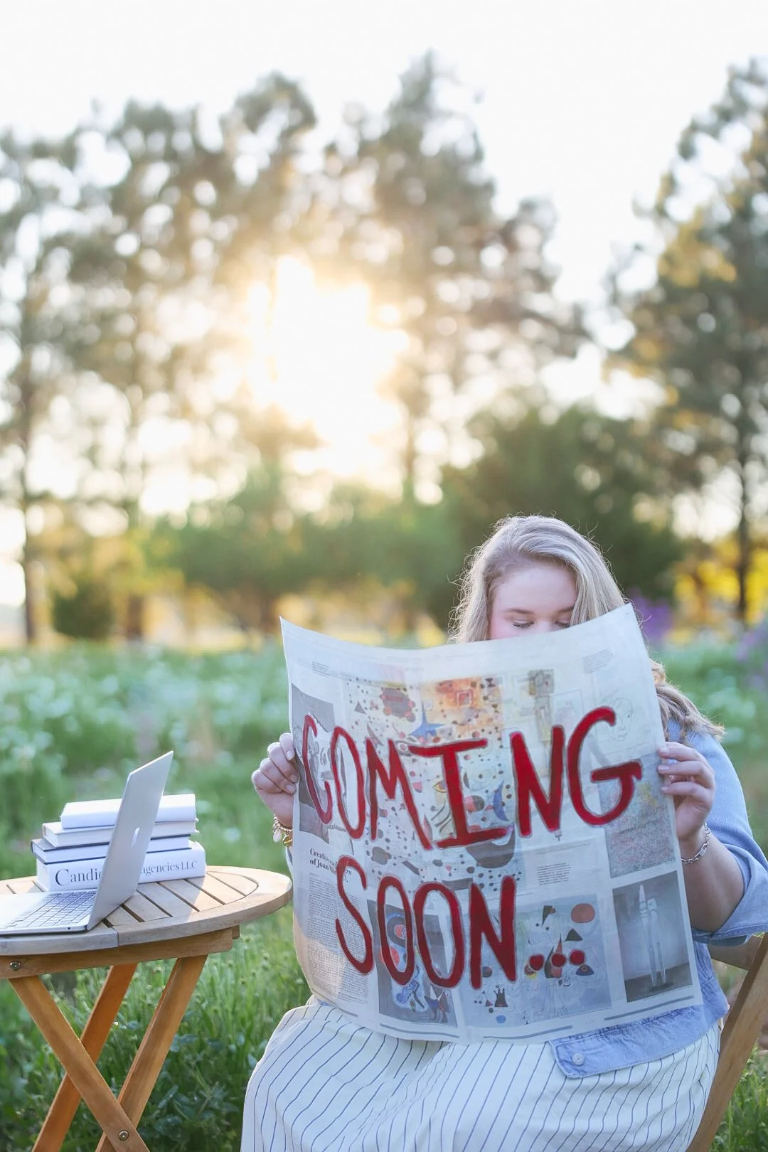 A woman sitting outdoors at a small wooden table reading a newspaper with 'COMING SOON' written in red ink. The background features trees and sunlight. On the table are a silver laptop and several books.