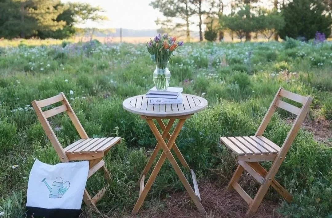 A small wooden round table with a glass vase of tulips and books, flanked by two wooden folding chairs, set outdoors in a grassy field with trees in the background.