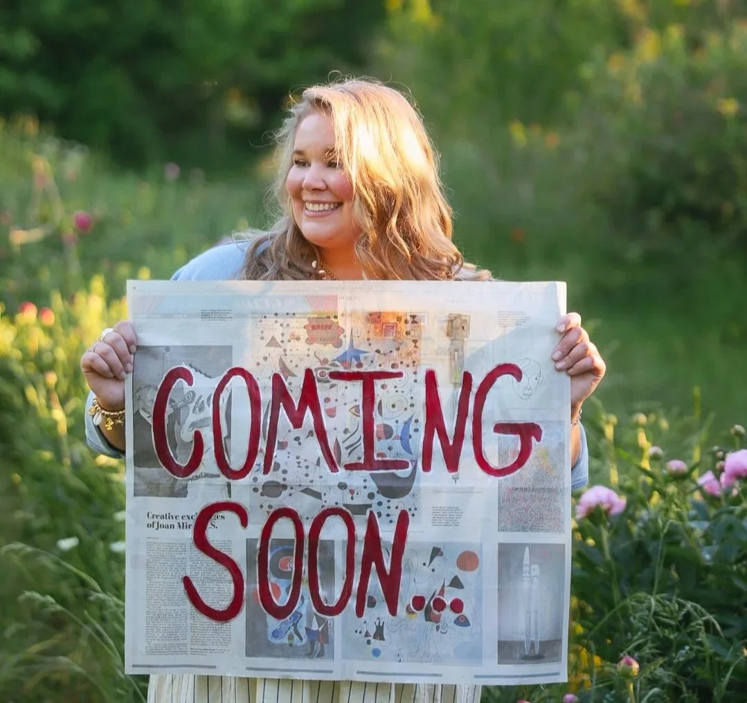 A woman with long wavy hair smiling while holding a sign that says "Coming Soon" in an outdoor setting with greenery and flowers.