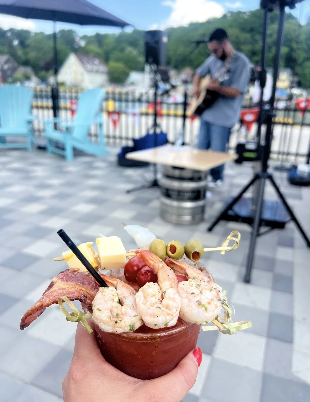 A person holding a cocktail garnished with shrimp, cheese, olives, cherry tomatoes, and bacon on toothpicks, with a musician playing guitar in the background at an outdoor venue.