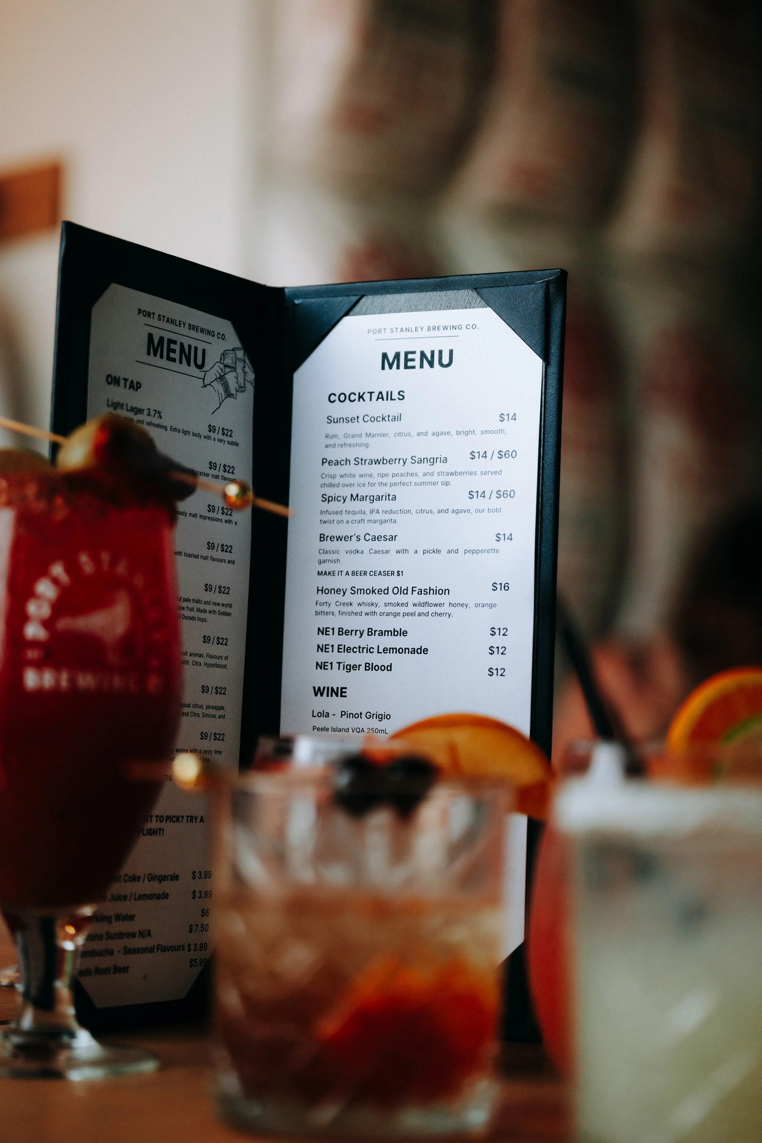 A black table menu with cocktail options at Port Stanley Brewing Co. surrounded by various colorful drinks with garnishes.
