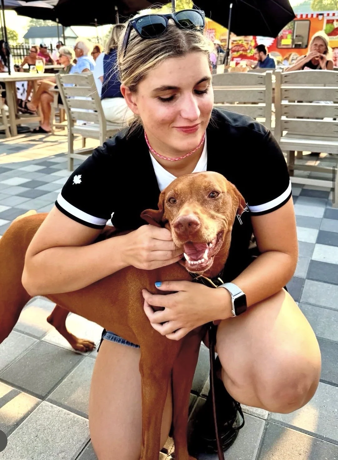 A woman with sunglasses on her head crouches down and hugs a happy, brown dog at an outdoor dining area. The background shows other people sitting at tables and food trucks.