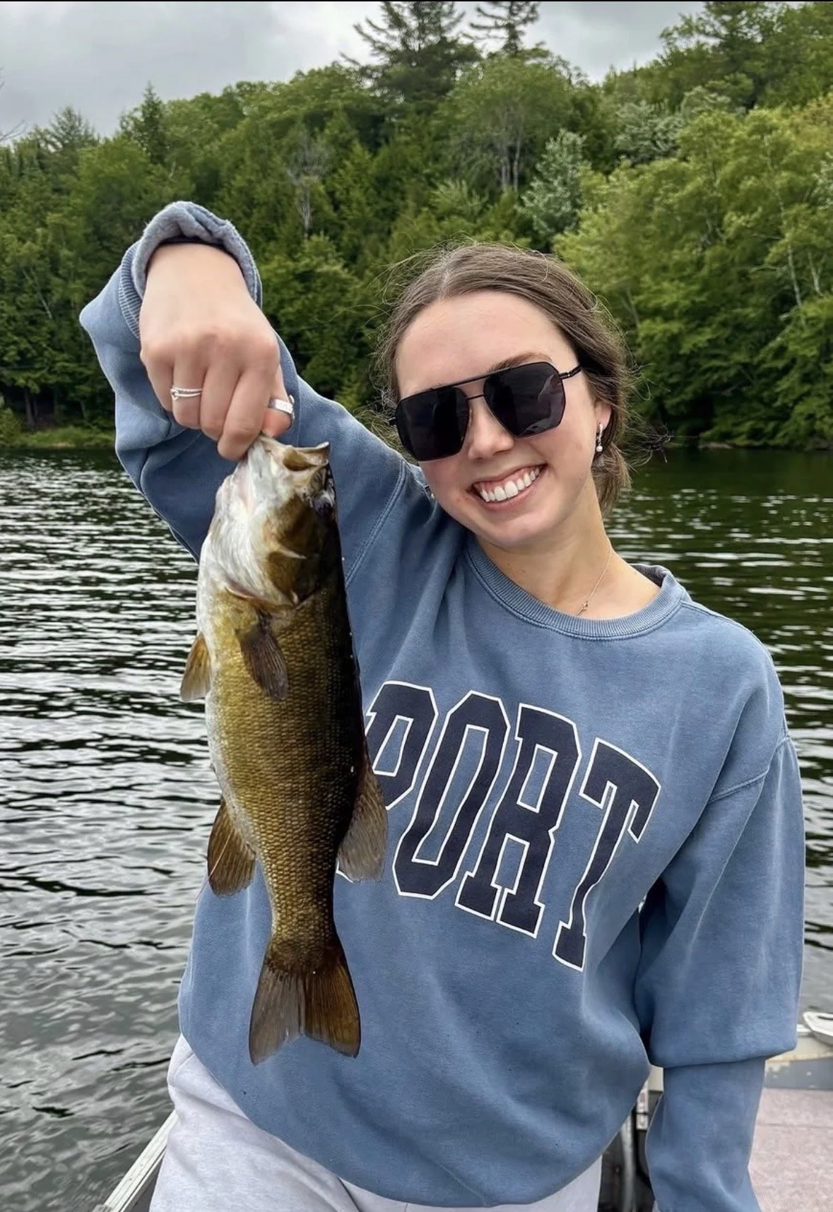 Smiling woman with sunglasses holding a freshly caught fish, standing on a boat on a lake with a forested hillside in the background.