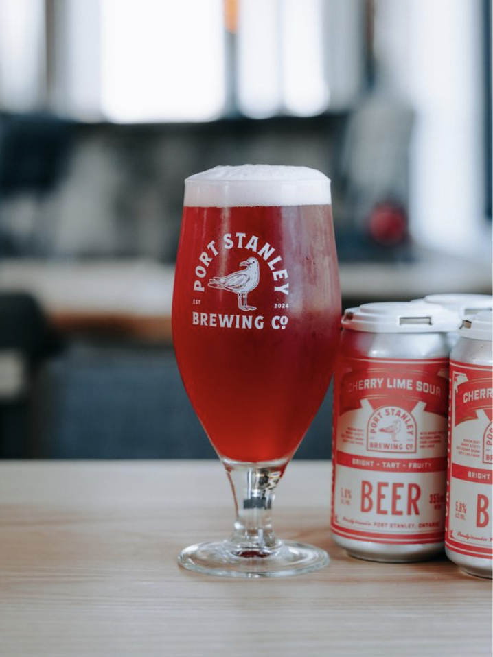 A glass of red beer with a foamy head on a wooden table, branded with Port Stanley Brewing Co, alongside cans of cherry lime sour beer.