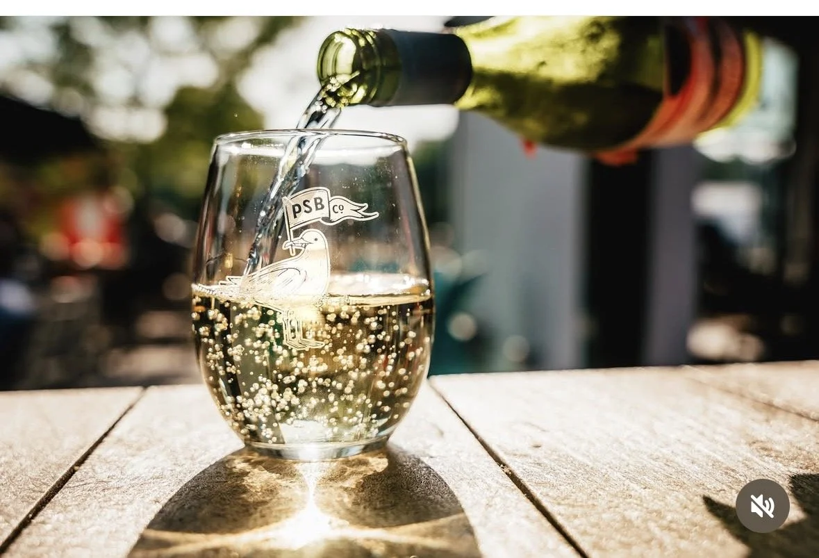 A glass of white wine being poured from a green bottle, with bubbles rising in the wine, on a wooden surface outdoors.