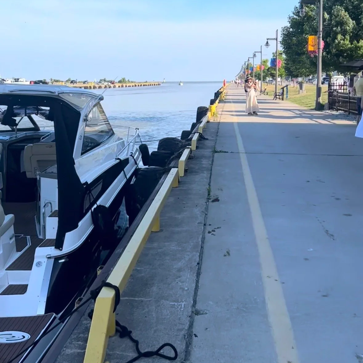 A marina promenade with boats docked on the water on the left and a walking path on the right. People are walking along the waterfront, with colorful banners hanging from street lamps and trees lining the sidewalk.