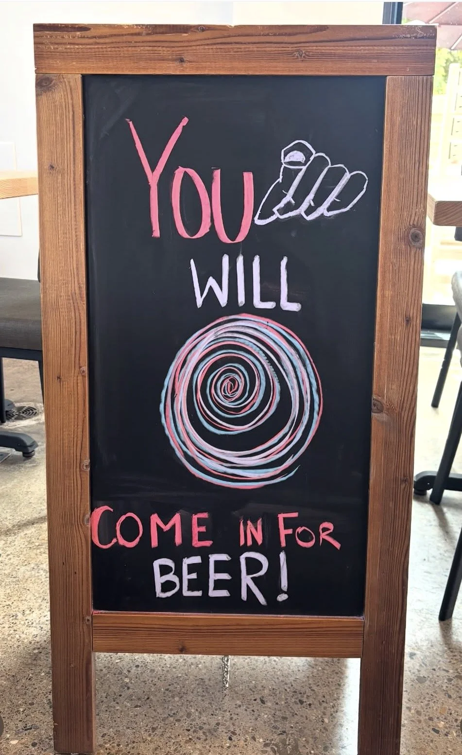 A black chalkboard sign with a wooden frame, displaying colorful hand-drawn text and images. The sign says, "You will come in for beer!" with the words "You" and "Come in for" written in pink, "Will" and "Beer!" in light purple. There is a spiral dra