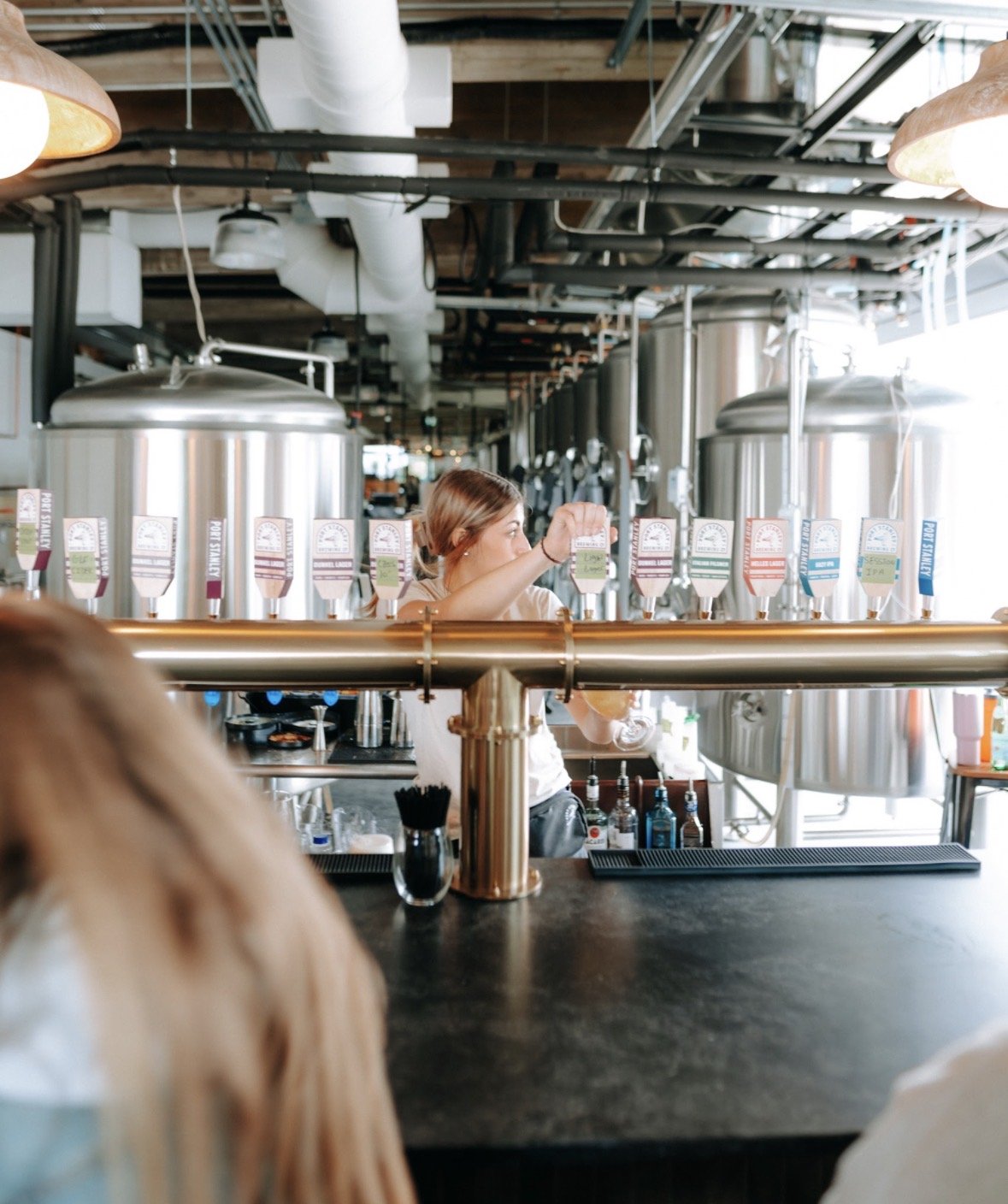 A woman working behind the bar at a brewery, pouring a beer from a tap system with multiple taps, large stainless steel fermentation tanks in the background, and a bar counter in the foreground.