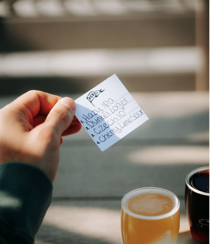 Person holding a small handwritten note with a list, next to a glass of beer and a dark beverage on a wooden table outdoors.