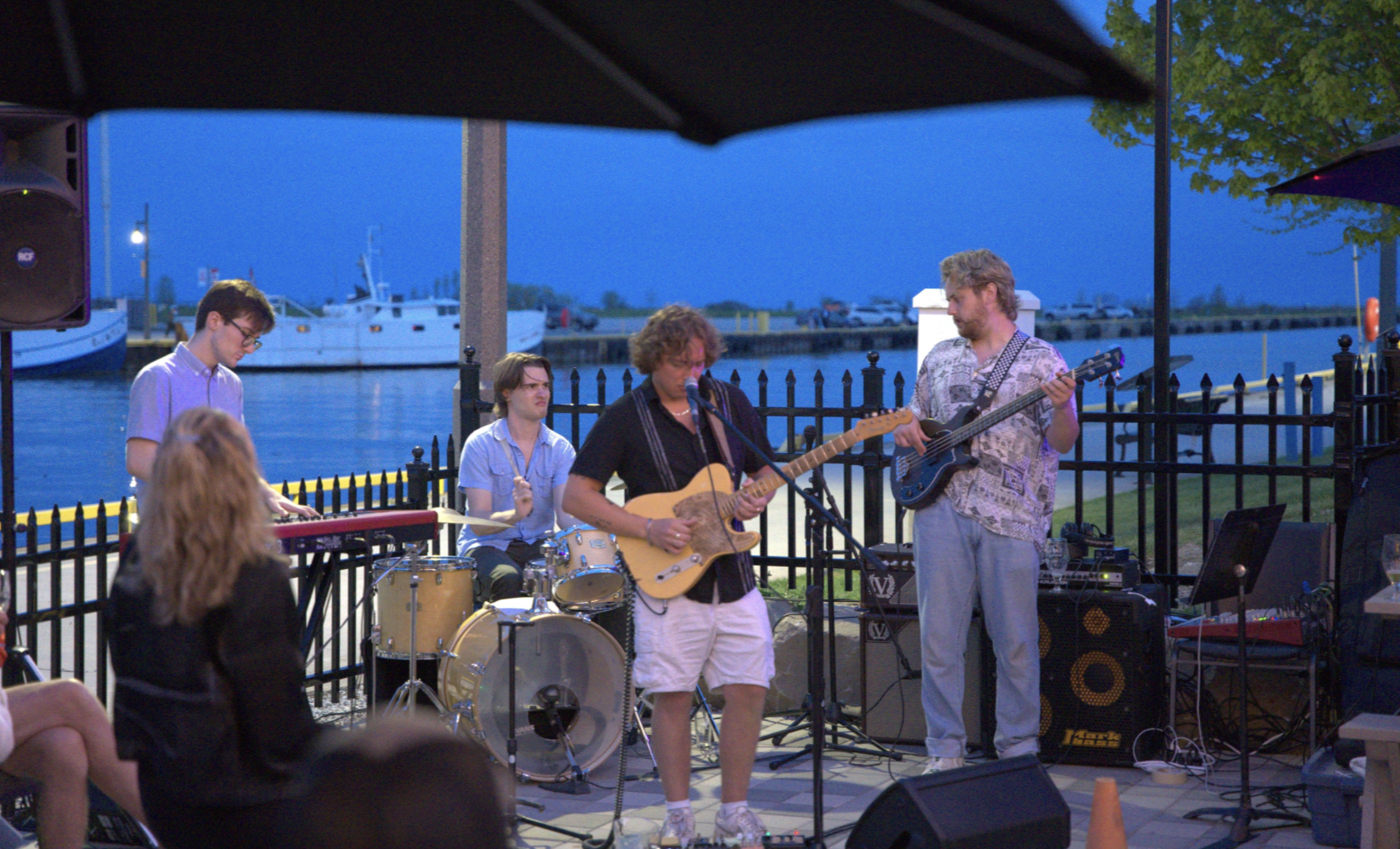 A band performs outdoors near a body of water at dusk, with four musicians playing instruments including drums, keyboard, and guitars, while audience members watch.