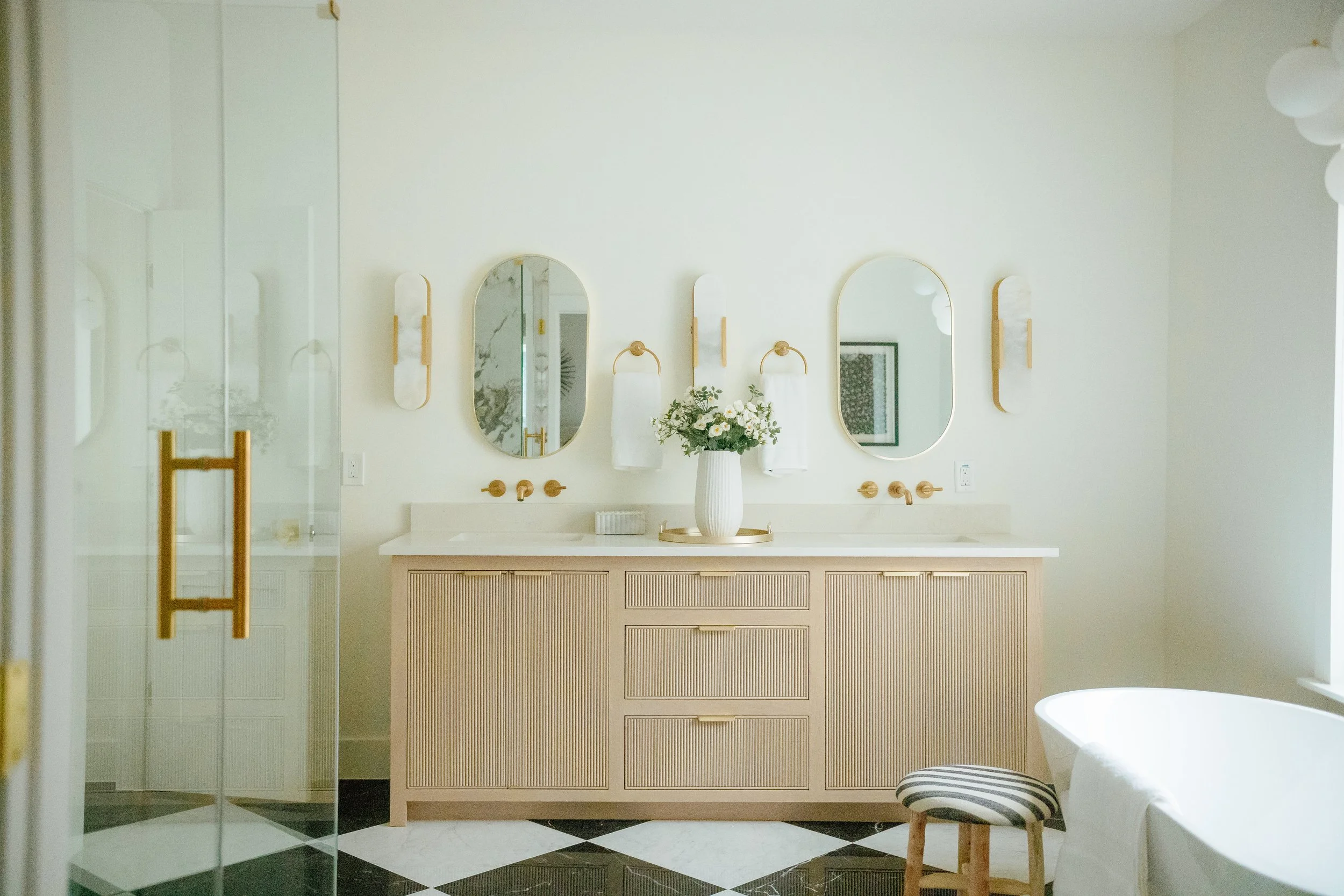 Modern bathroom with dual sinks, oval mirrors, and white walls, featuring a vase of flowers on the counter, a striped stool, and a partial view of a bathtub. New construction. Bathroom renovation.