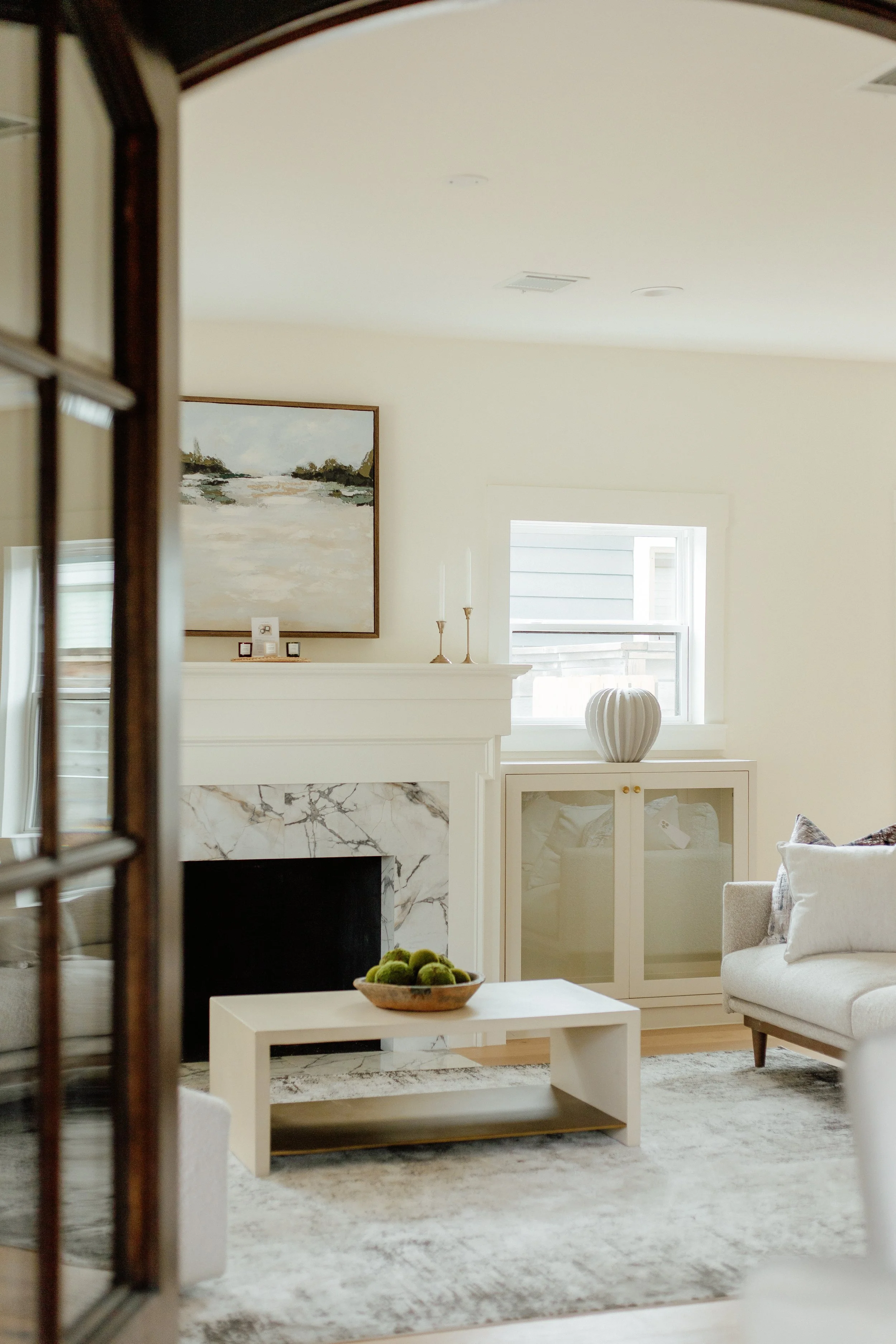 Living room with white walls, a white marble fireplace, a large painting above the mantel, a window with white trim, a beige sofa with pillows, and a white coffee table with a bowl of green decorative balls. Living room transformation.