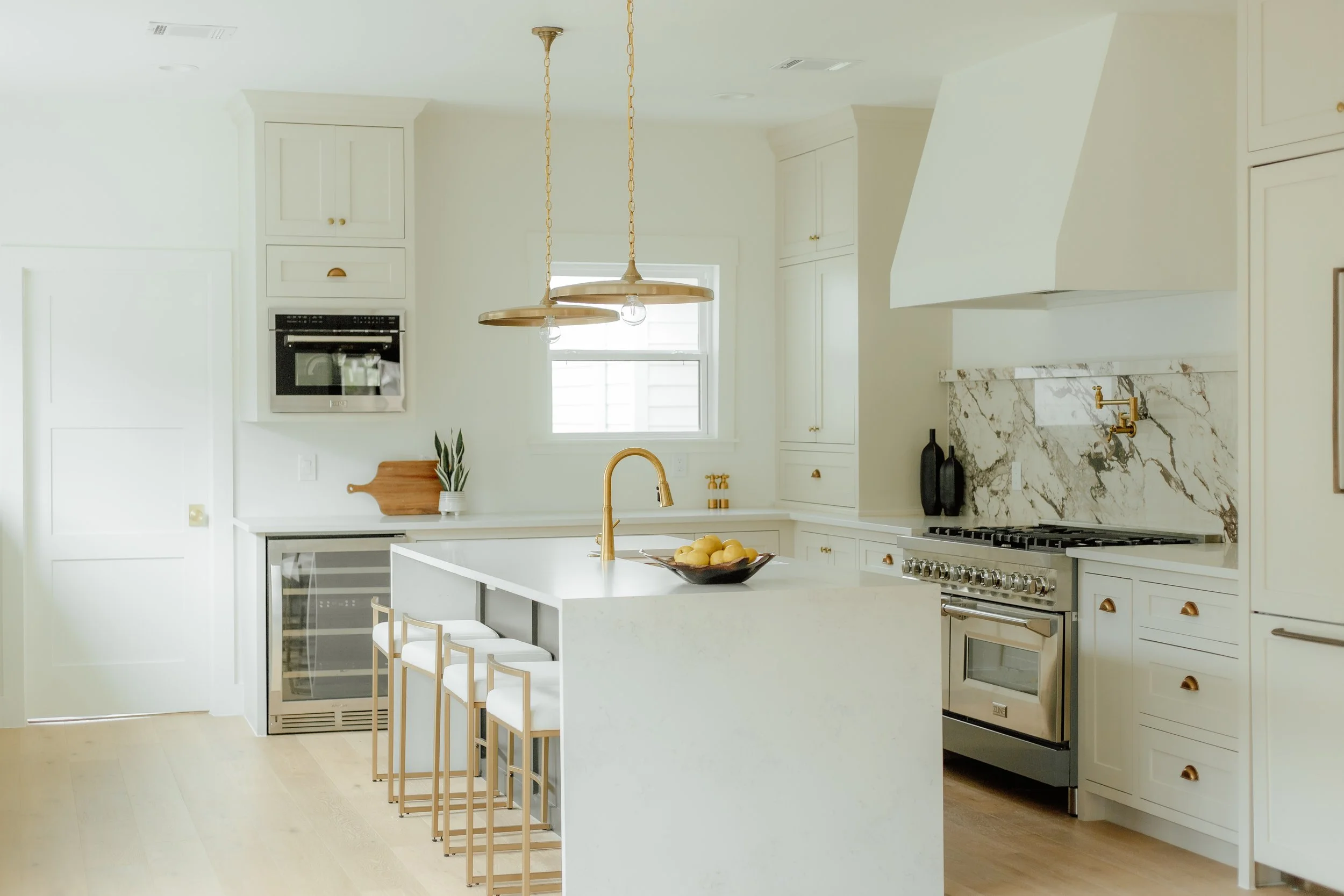 Bright, modern kitchen with white cabinets, marble backsplash, stainless steel oven, and kitchen island with gold faucet and four white bar stools with gold legs.