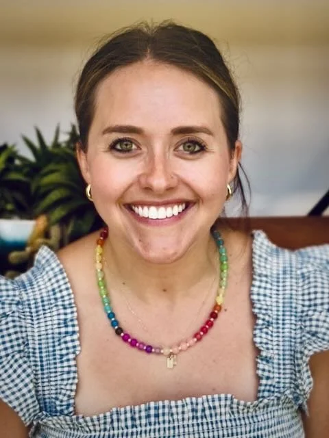 A woman with brown hair tied back, smiling, wearing a navy blue shirt and hoop earrings. Behind her are green vines with small leaves, and a bowl of food is partially visible on the left.