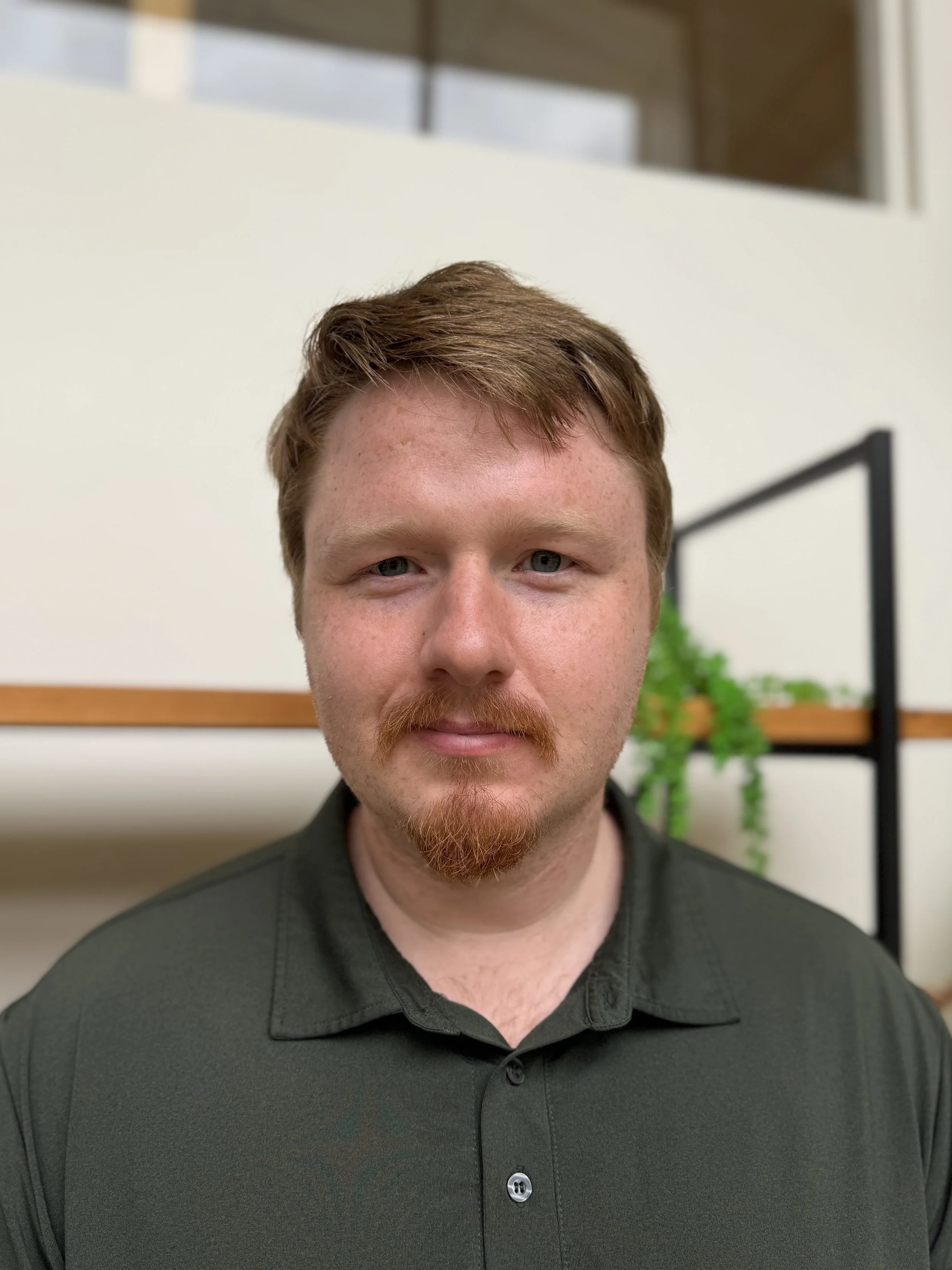 A smiling man with short red hair and a green button-up shirt, standing indoors with green houseplants and a wood shelf in the background.