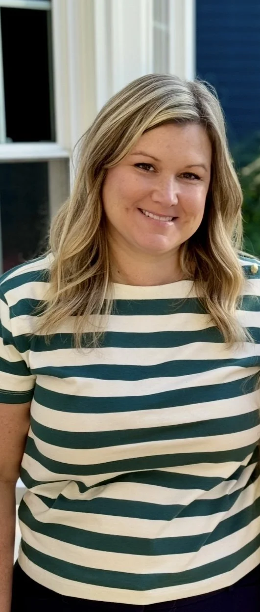 A woman with blonde hair wearing a green and white striped shirt, smiling, standing outdoors near a window with a blue house in the background.