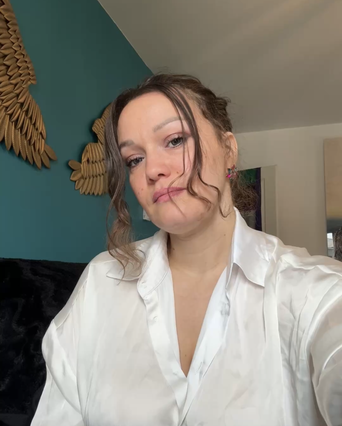 Une femme avec des cheveux bruns ondulés, portant une chemise blanche, prend une selfie dans une pièce avec un mur vert en arrière-plan et une décoration en bois.