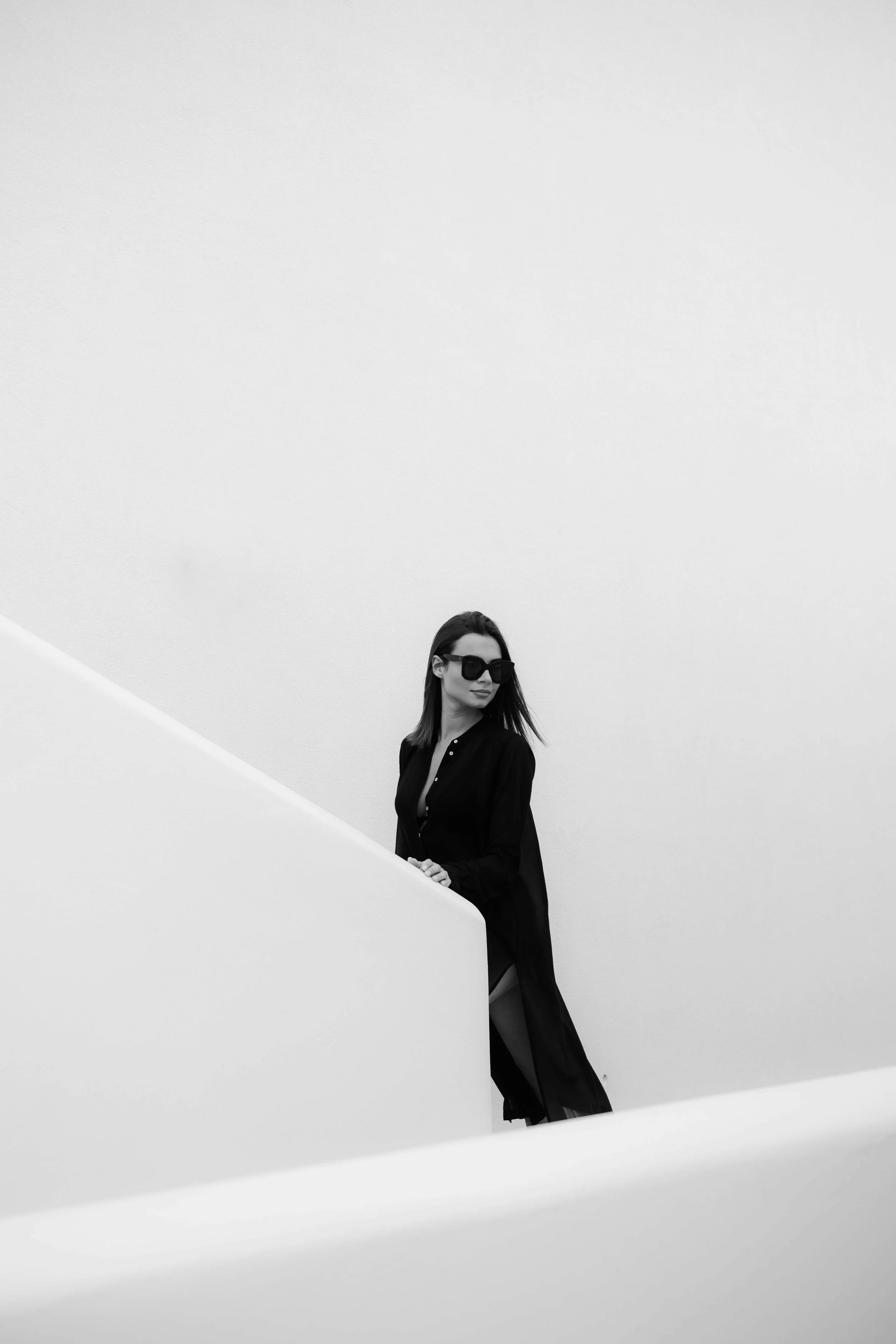 A woman wearing dark sunglasses and a long black dress standing on a staircase against a plain wall.