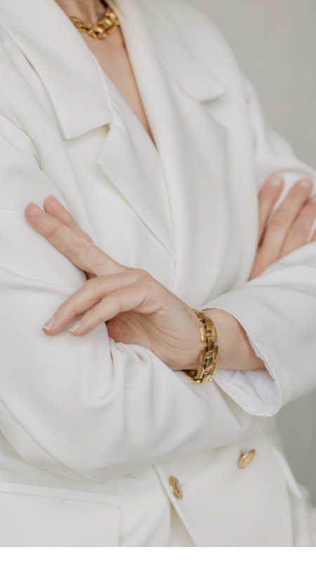 Nahaufnahme einer Person in einem weißen Blazer mit verschränkten Armen, goldene Armbanduhr und Halskette.