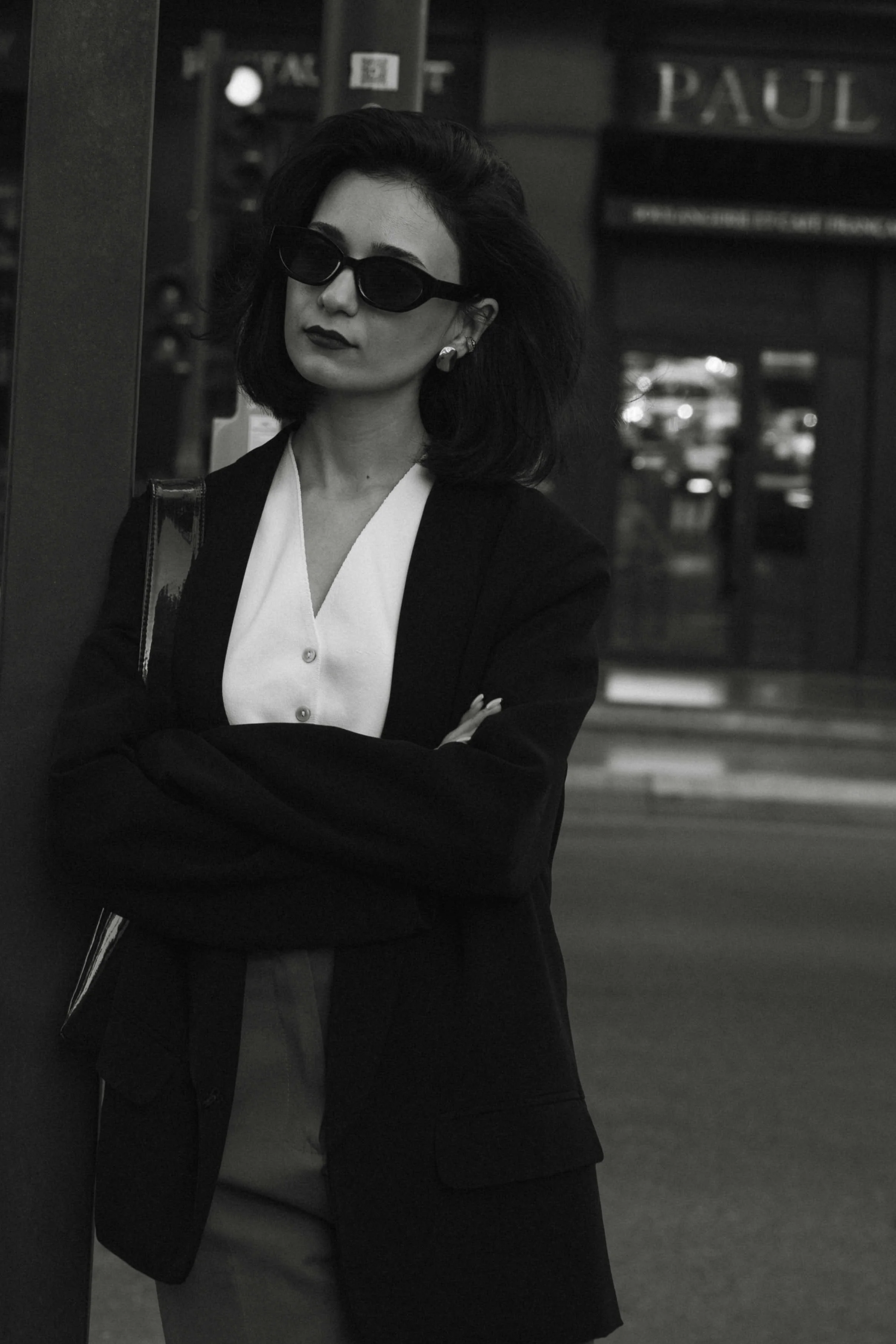 A woman with dark hair, wearing sunglasses, a white blouse, and a black blazer, stands with her arms crossed in front of a building with outdoor signage, in black and white photography.