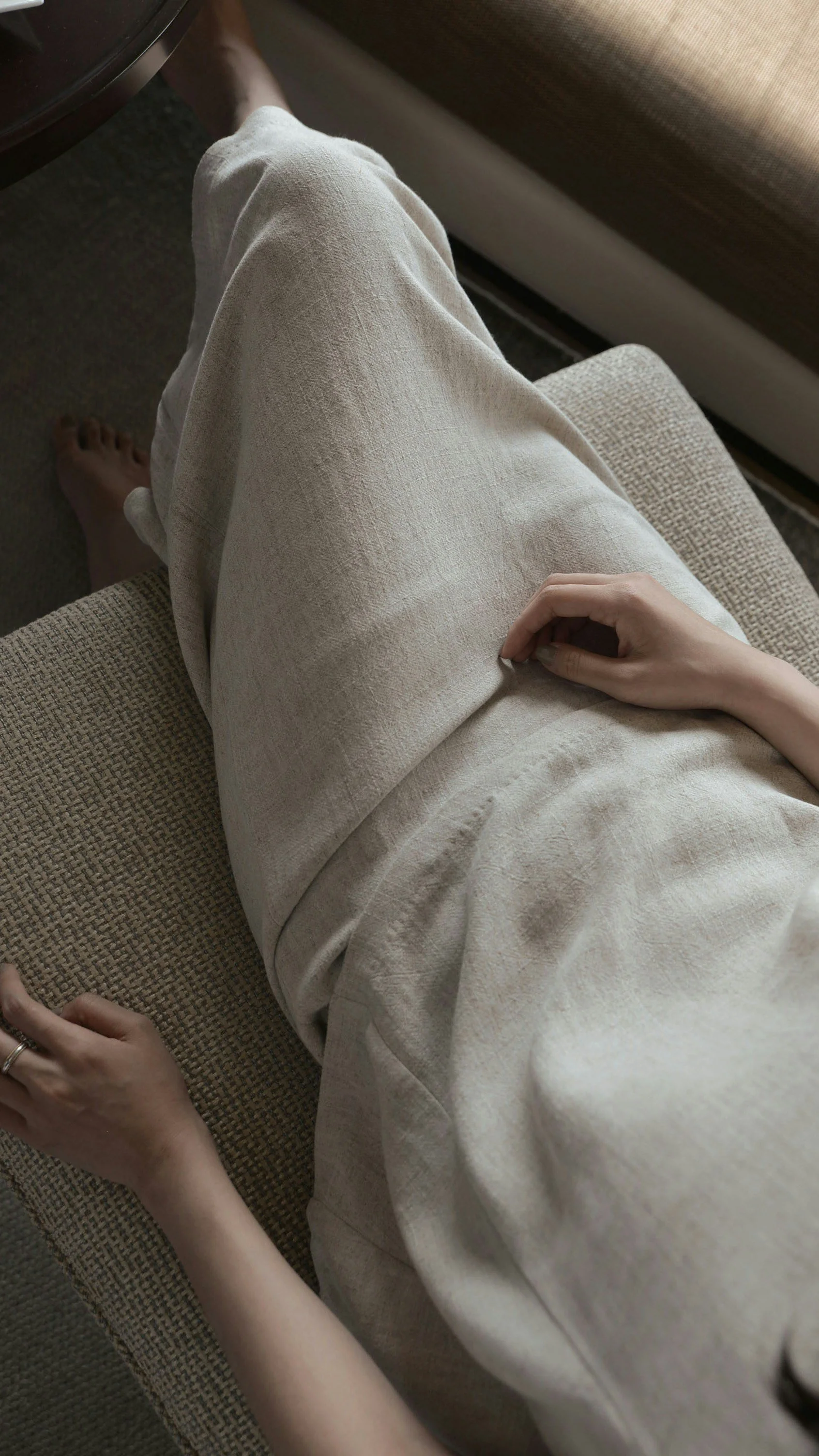 Person sitting on a beige fabric sofa with beige pants and a beige shirt, resting hands on lap, with one foot visible on the carpeted floor.