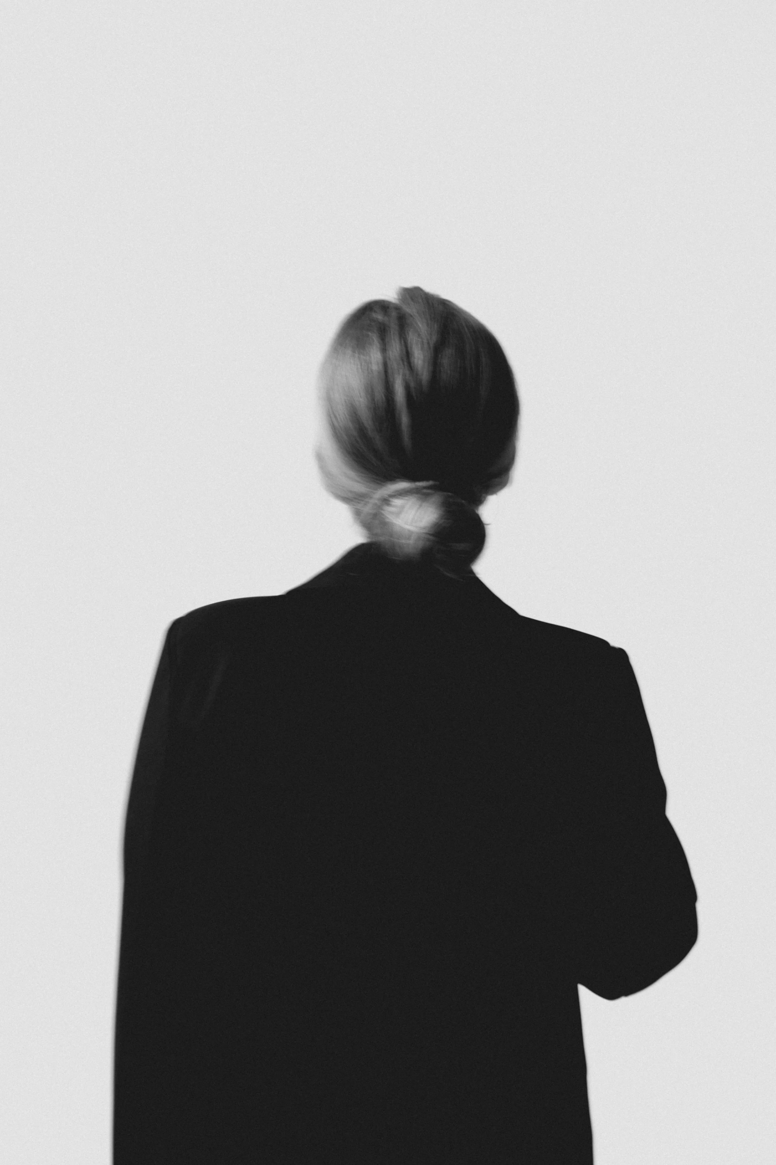 Back view of a woman with her hair in a bun, wearing a dark blazer, standing against a plain, light background.