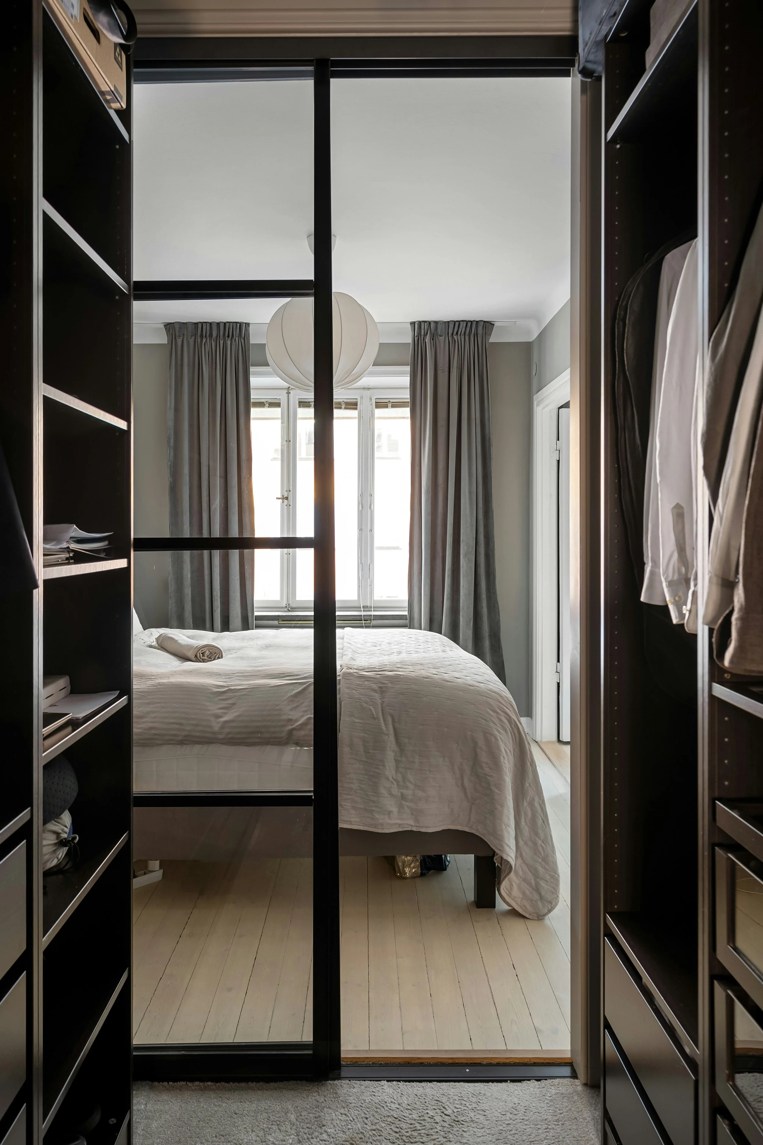 A bedroom seen through sliding mirror closet doors, featuring a bed with white bedding, a window with closed curtains, and a ceiling light fixture.