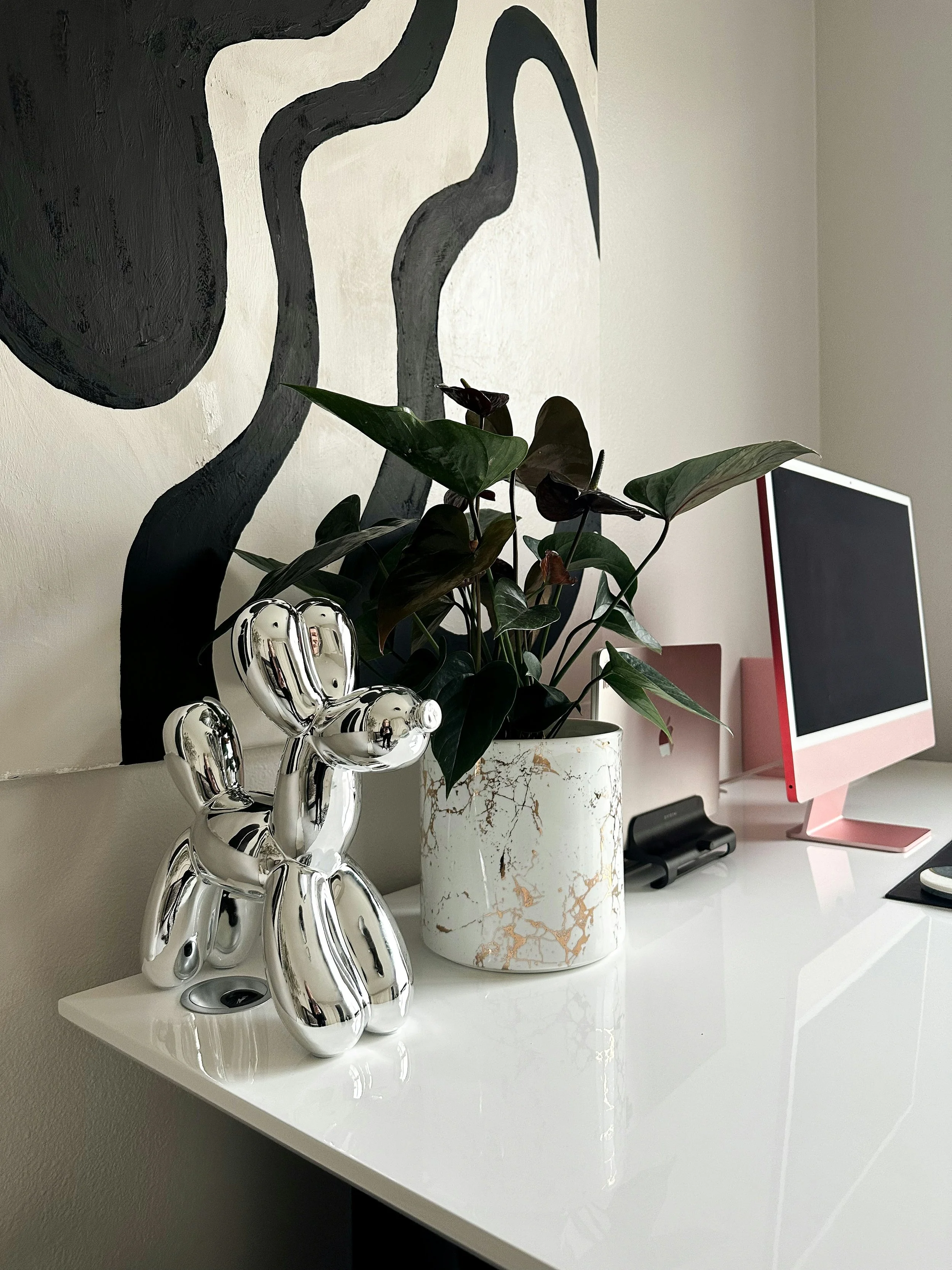 Decorative desk setup with a silver balloon dog sculpture, a potted plant with dark green and brown leaves, a white marble-patterned planter, a computer monitor with a pink stand, and a black remote control on a white glossy desk against a wall with