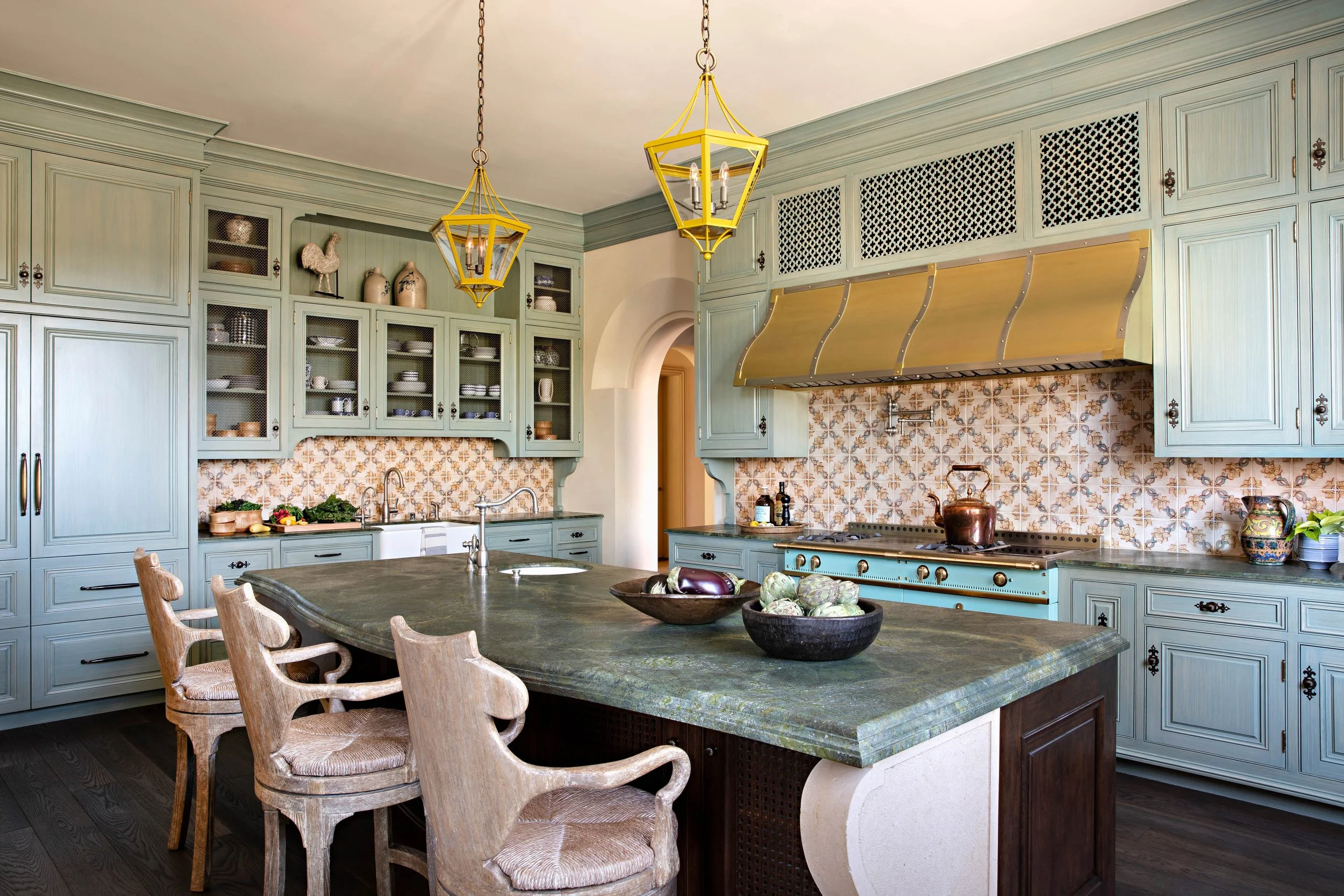 Kitchen with pastel green cabinets, a large kitchen island with a dark countertop, and a vintage-style backsplash with floral tiles. Two yellow pendant lights hang above the island, and there are wooden chairs around it. The kitchen has decorative di